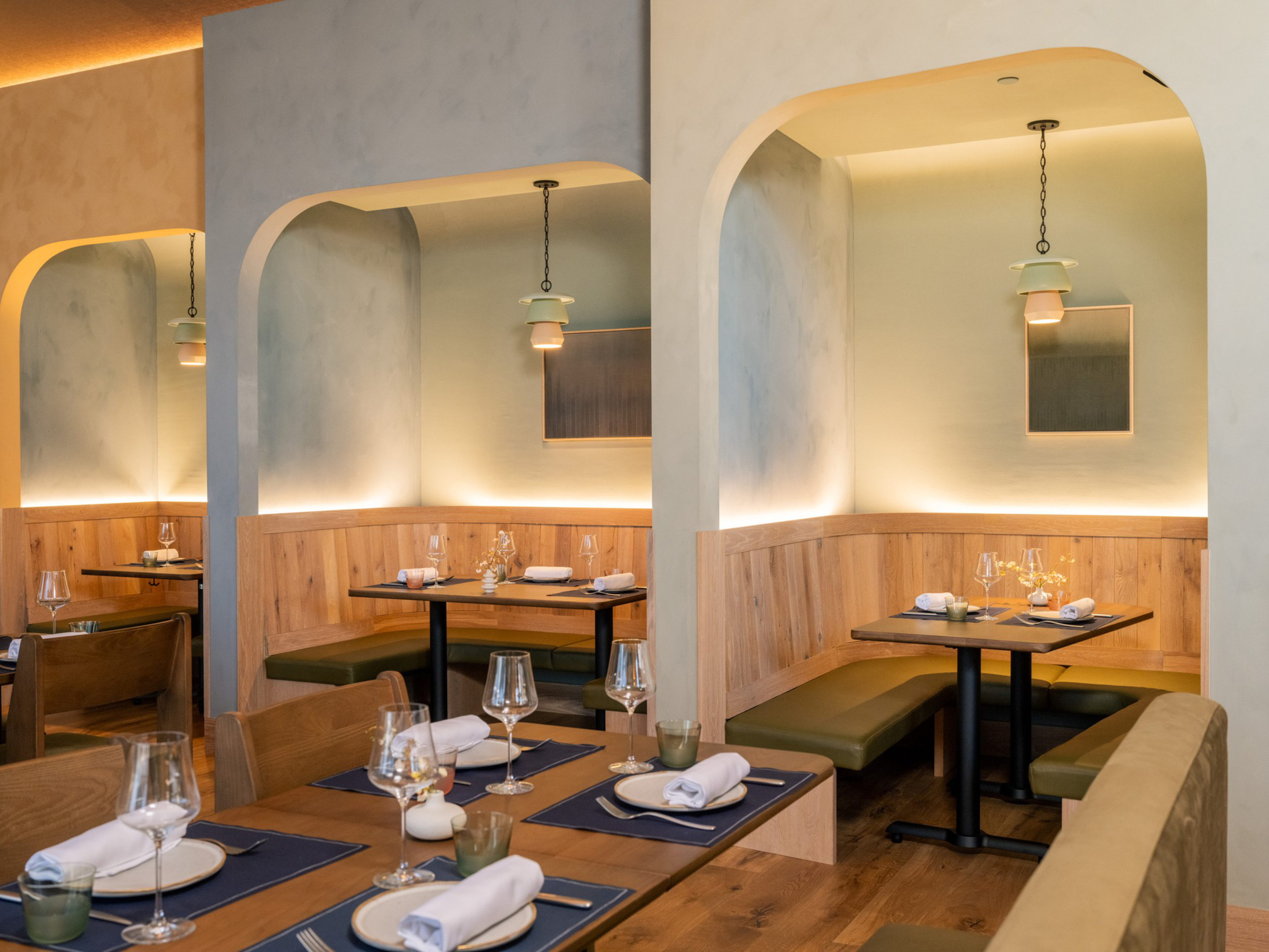 Cozy restaurant booths with wooden paneling and olive-green cushions sit under soft pendant lighting, surrounded by minimalist decor and set tables.