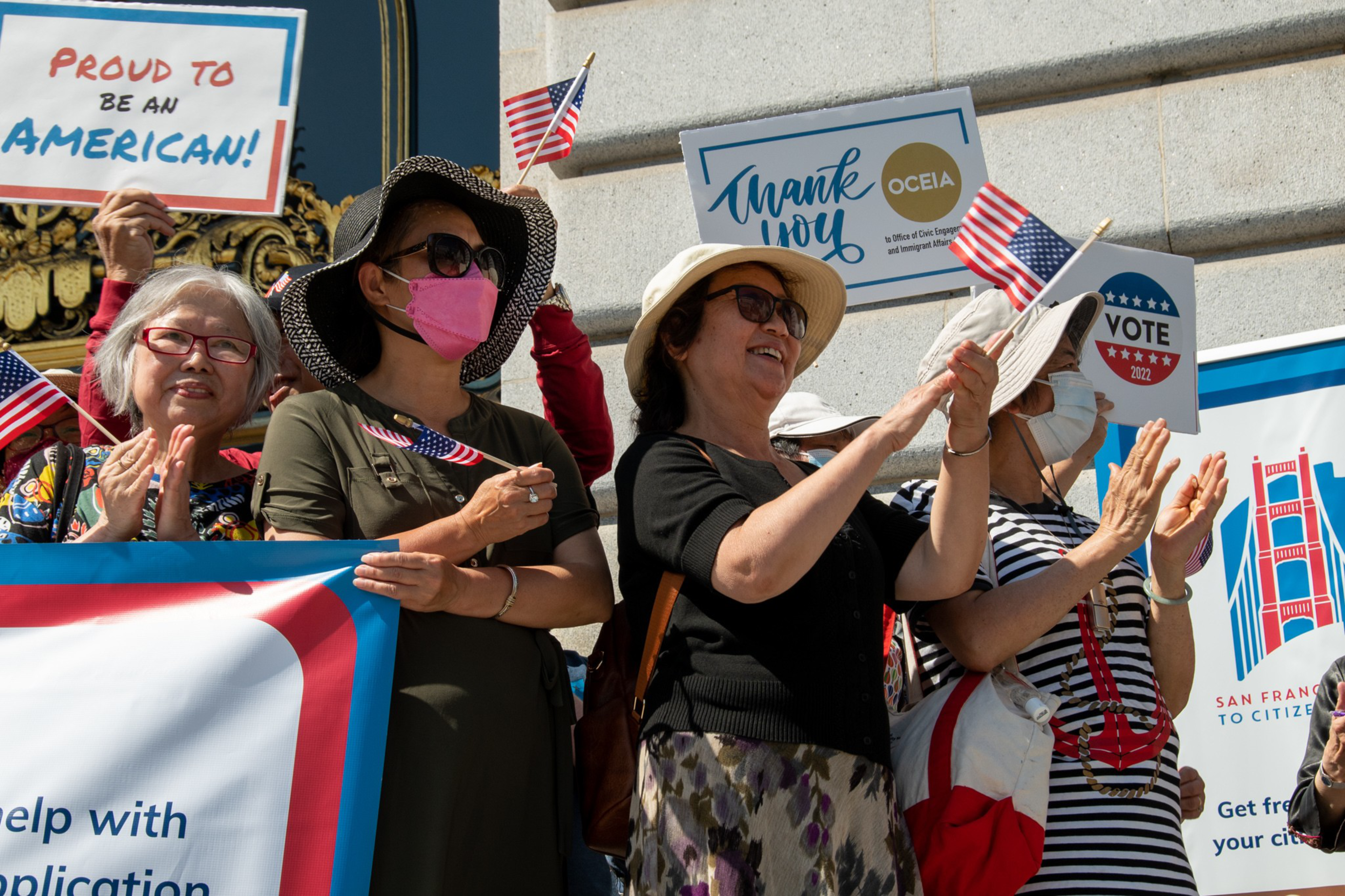 A group of diverse people hold American flags and signs with messages like "Proud to be an American!" and "Vote 2022" while clapping in sunlight.