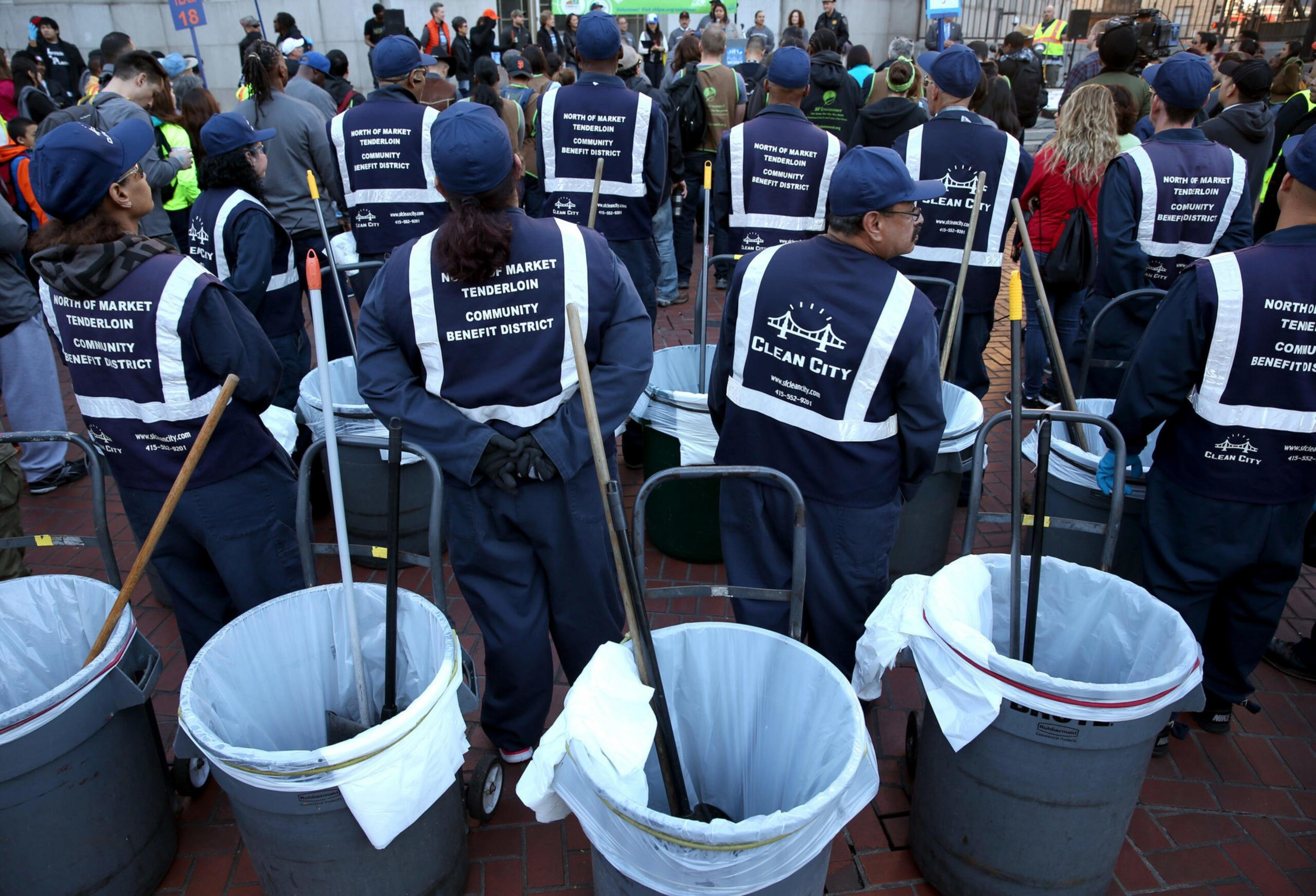 A group of sanitation workers in uniforms and vests labeled "Clean City" and "North of Market Tenderloin Community Benefit District" stand near trash cans with brooms inside.