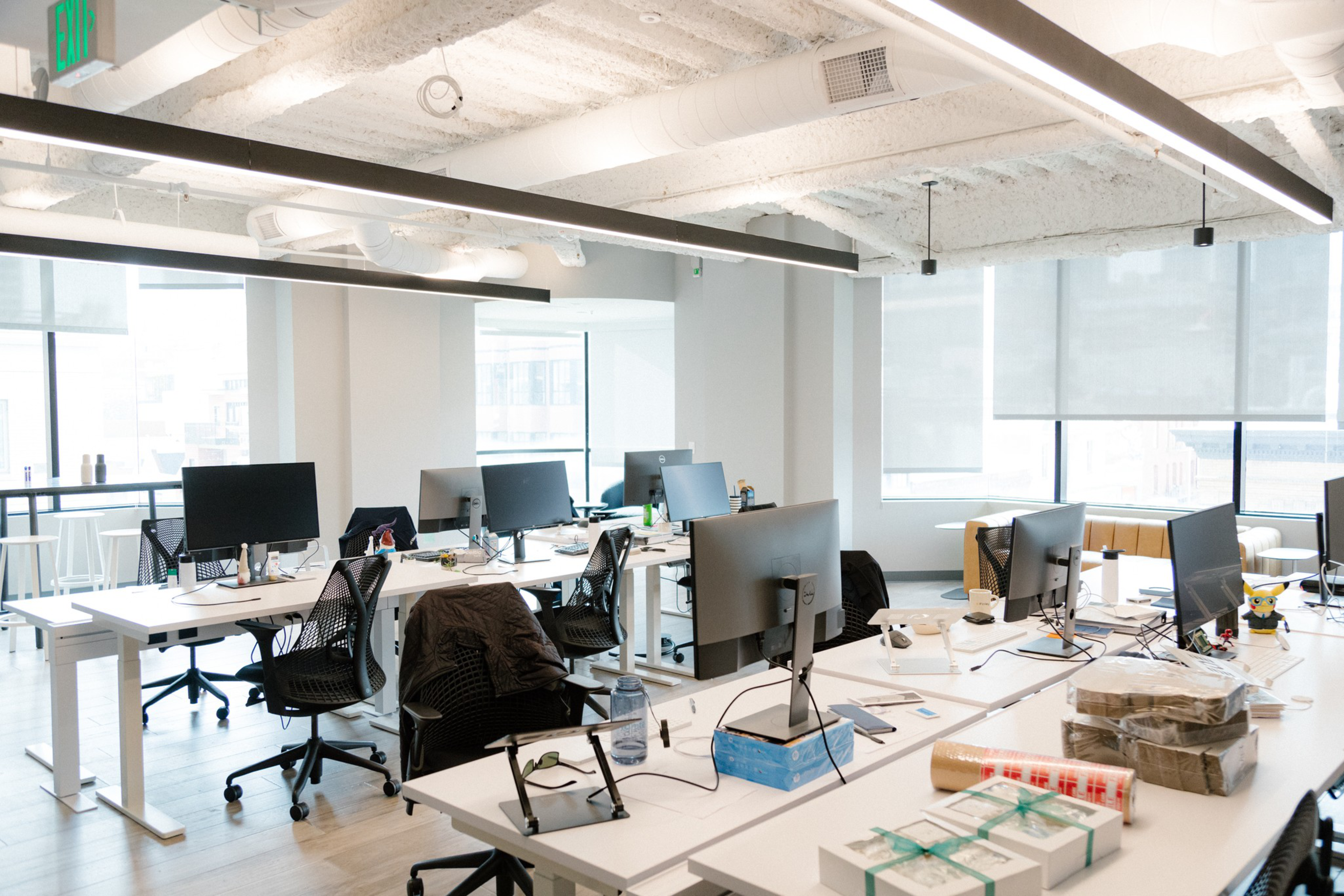 An open office space with multiple desks, computers, and chairs. There are large windows providing natural light and a high ceiling with exposed ductwork.
