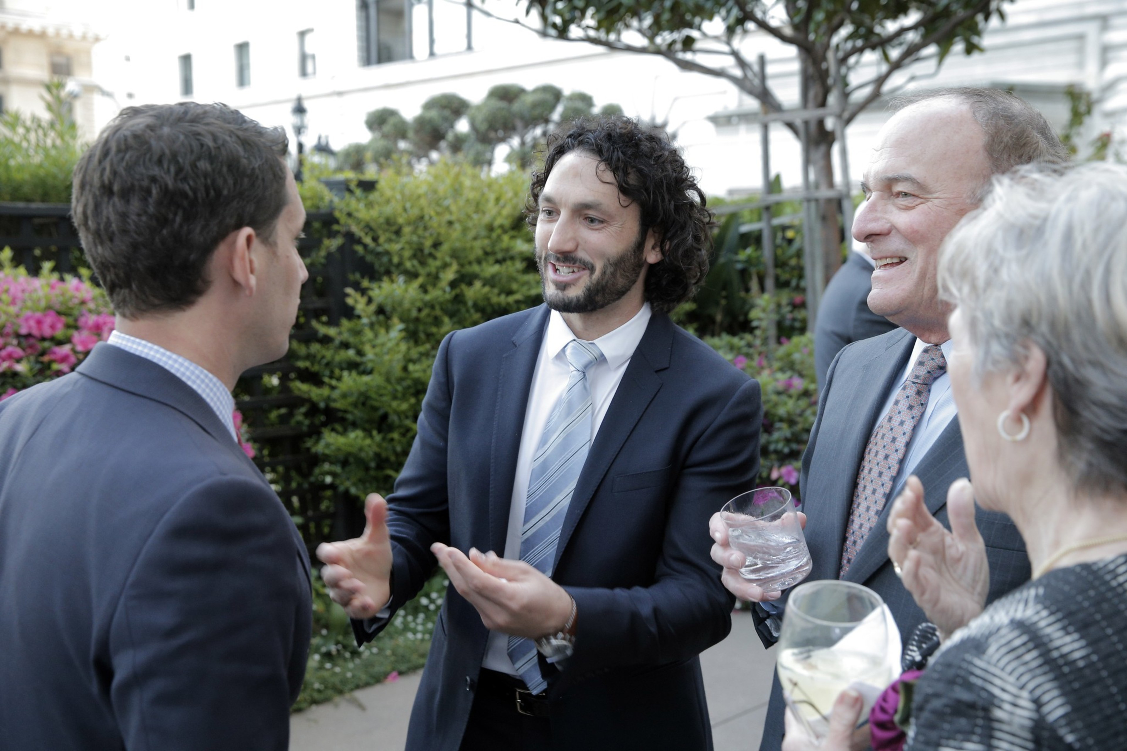 Four people in formal attire engage in conversation outdoors, holding drinks, with greenery and flowers in the background.