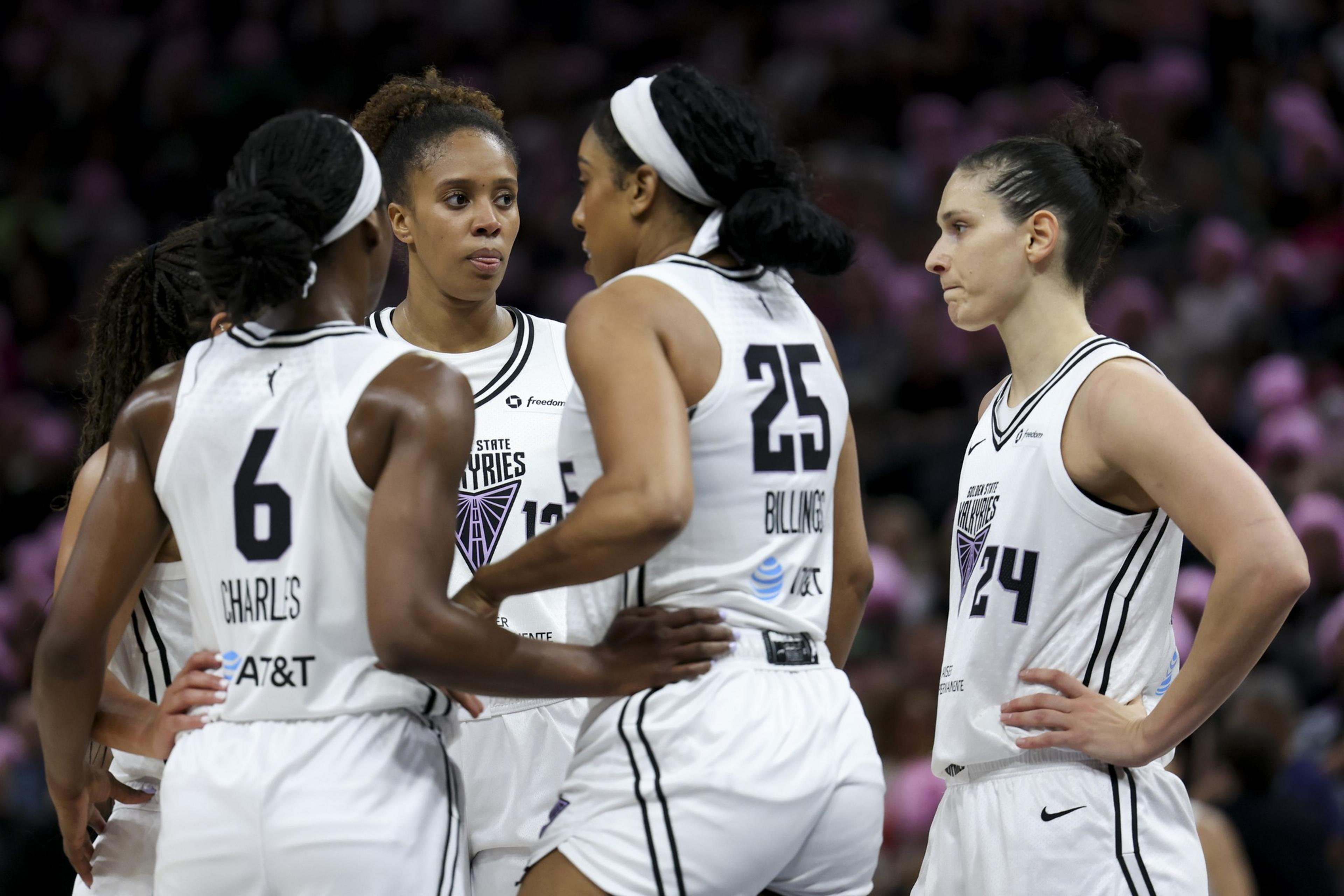 Four female basketball players in white uniforms stand closely, appearing focused and engaged in a team discussion during a game.