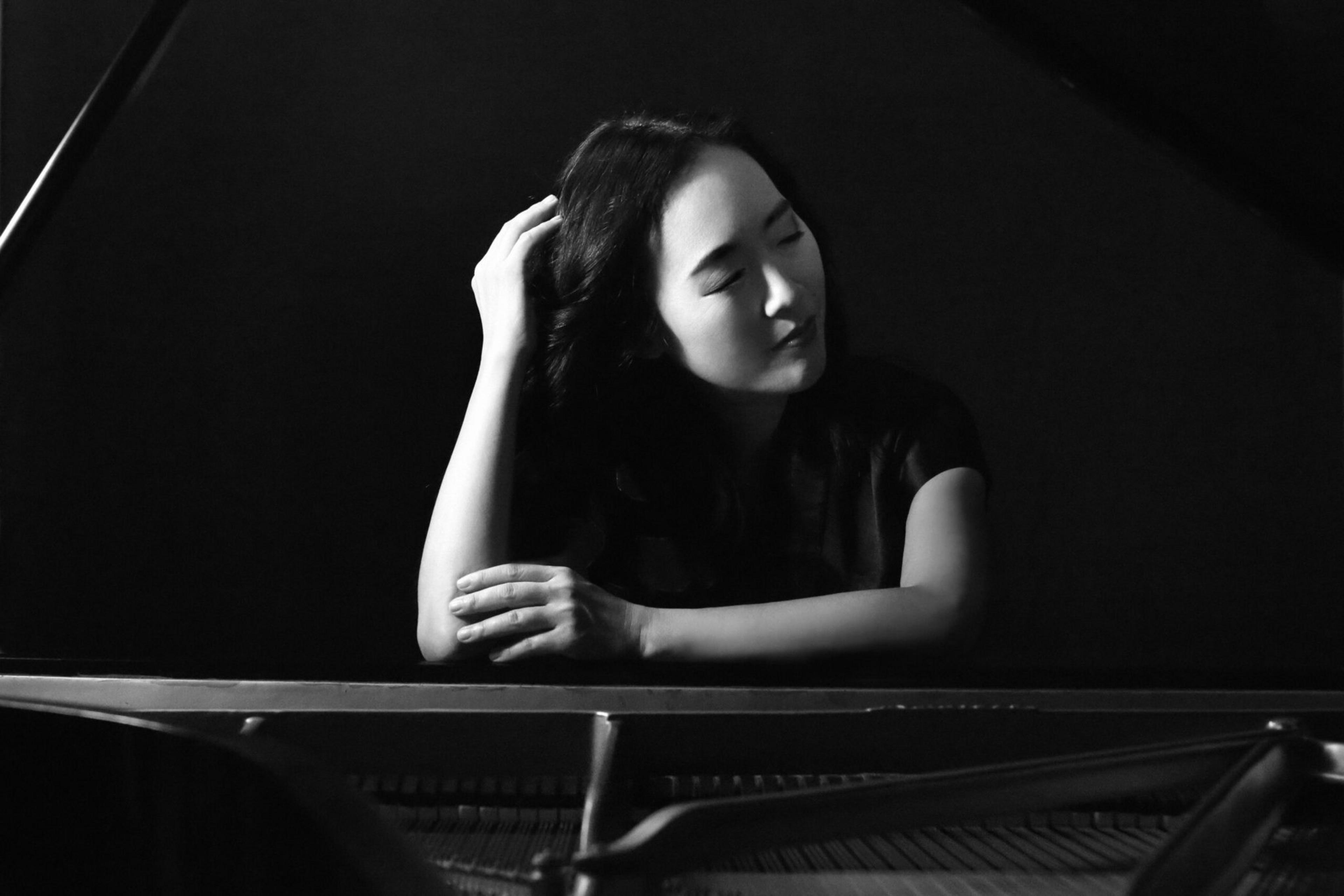 A woman with long dark hair rests her arms on a piano, her eyes closed and one hand touching her hair, against a dark background.