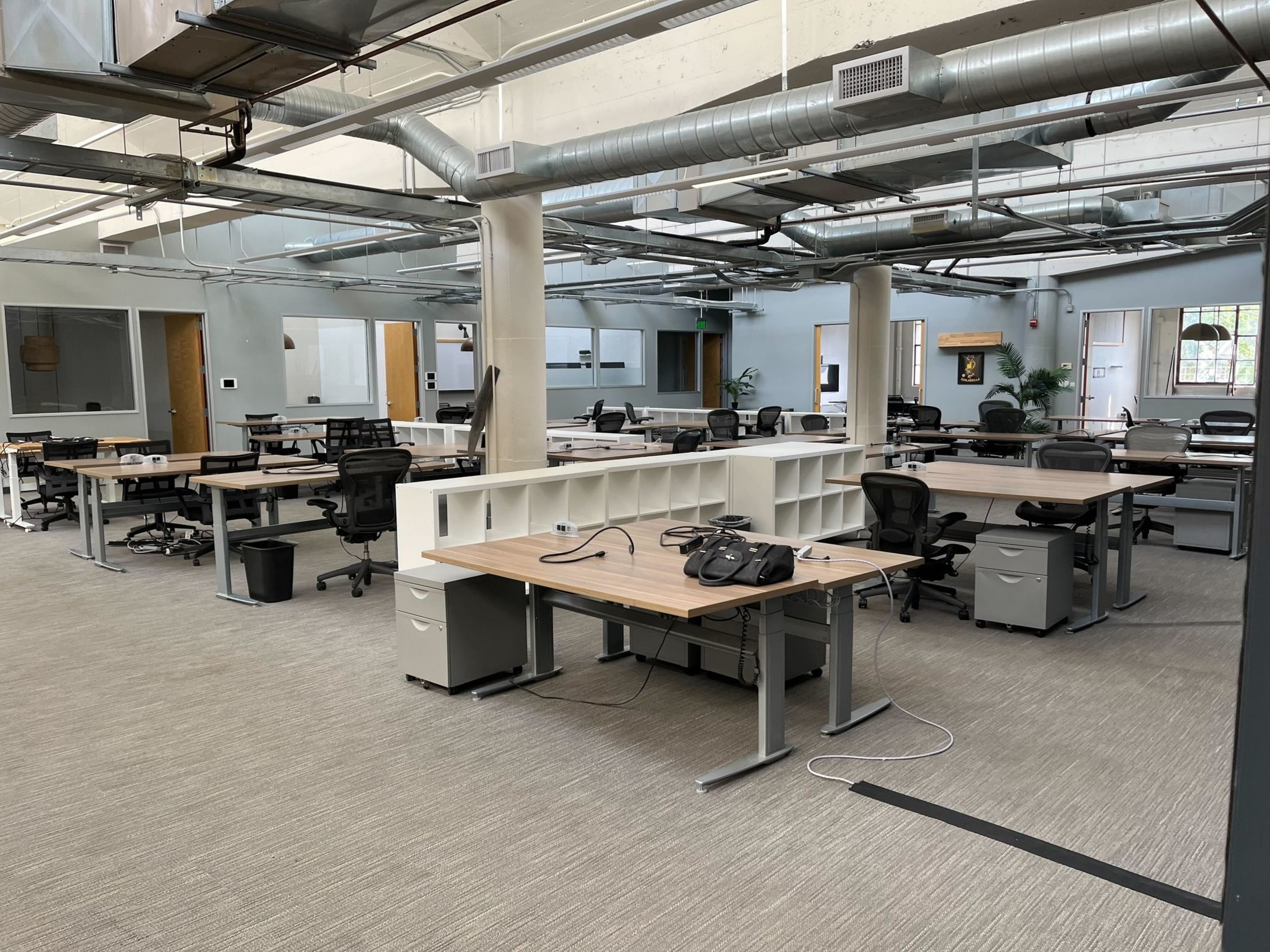The image shows a spacious, open-plan office with various empty desks, chairs, overhead ducts, and ceiling pipes, creating an industrial look.