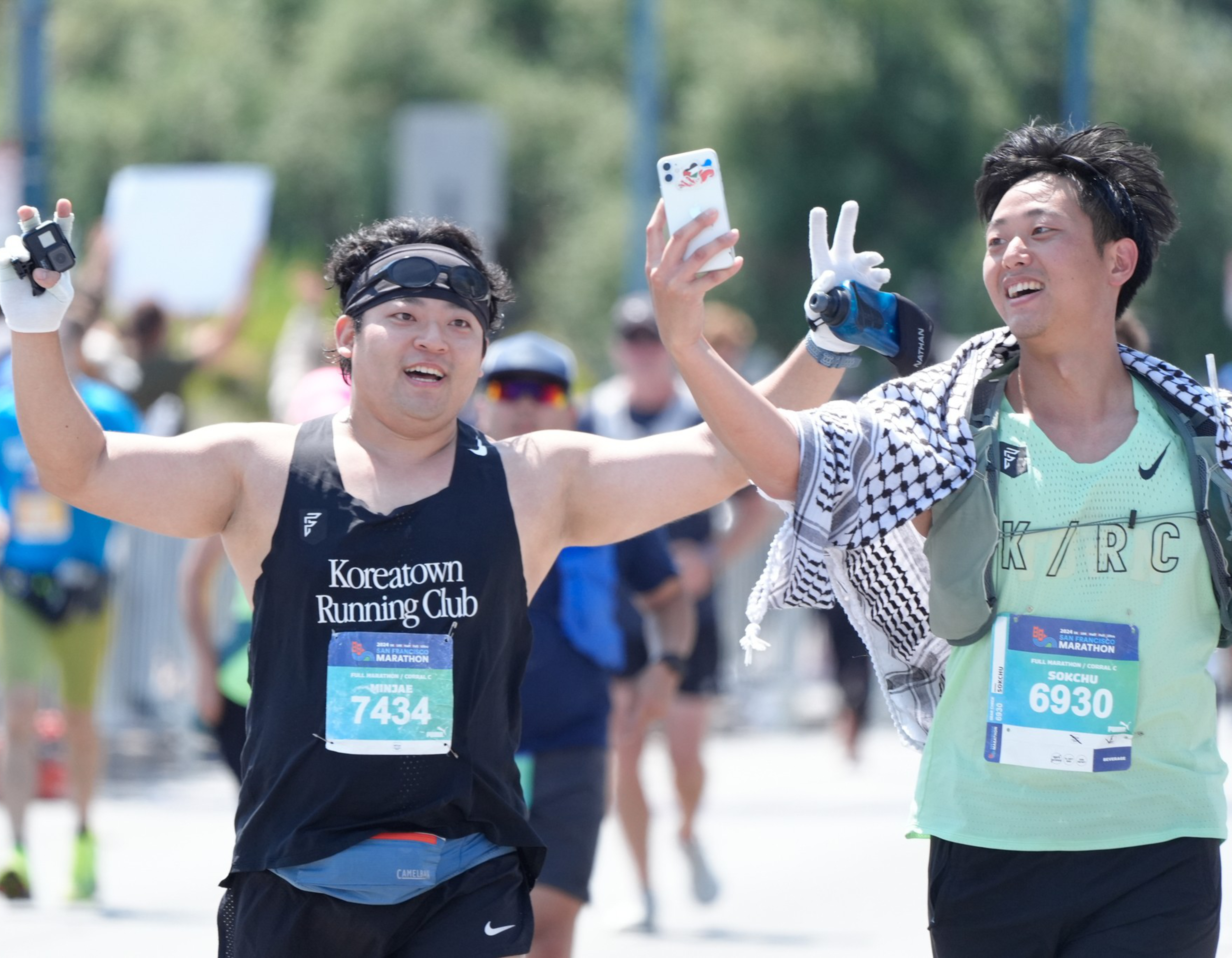 Two marathon runners, smiling and raising their hands, are taking a selfie. One wears a &quot;Koreatown Running Club&quot; shirt and the other has a &quot;K/R/C&quot; shirt and a striped scarf.