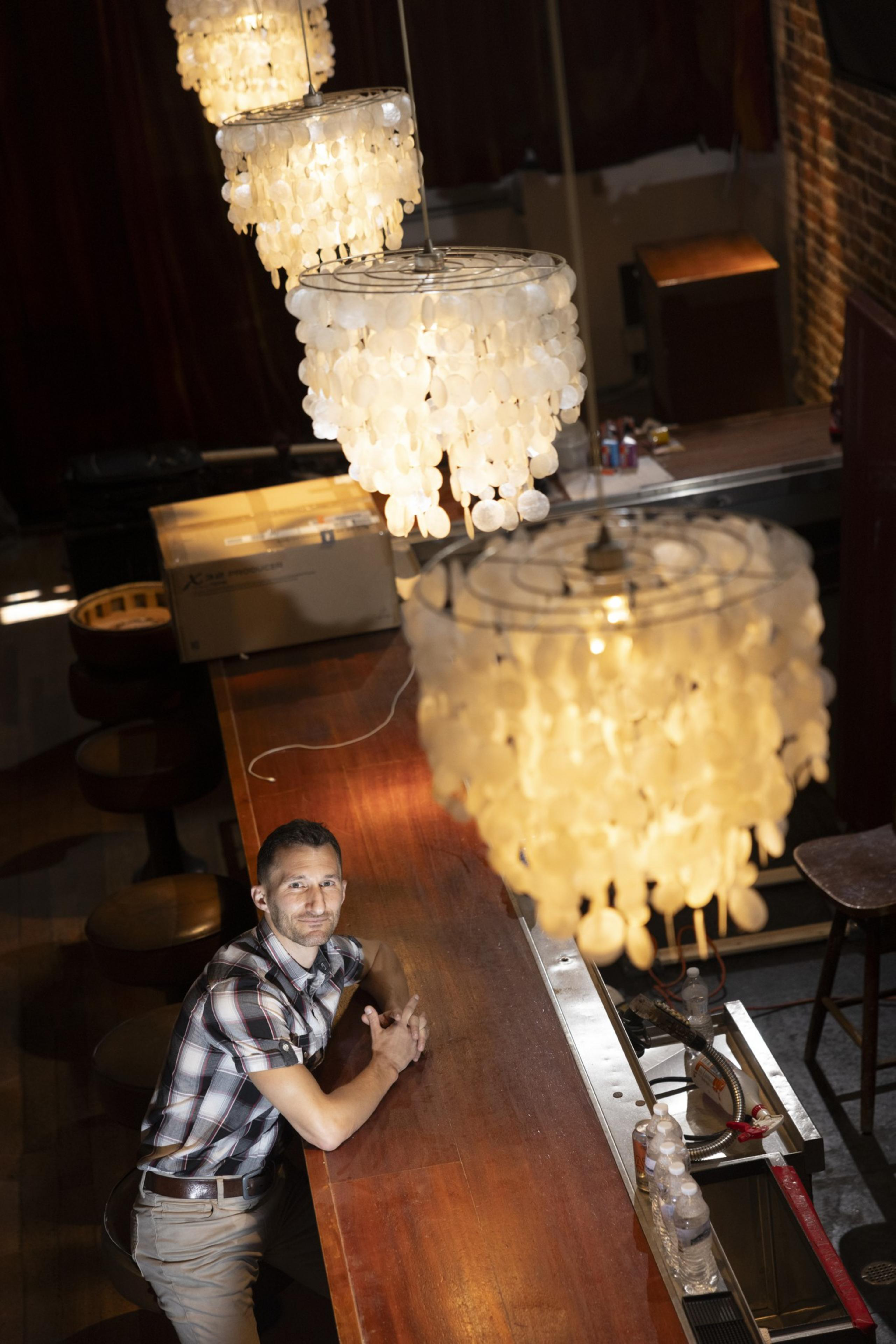 A man in a plaid shirt leans against a bar counter, illuminated by large, ornate chandeliers hanging overhead. The setting is cozy with a warm ambiance.