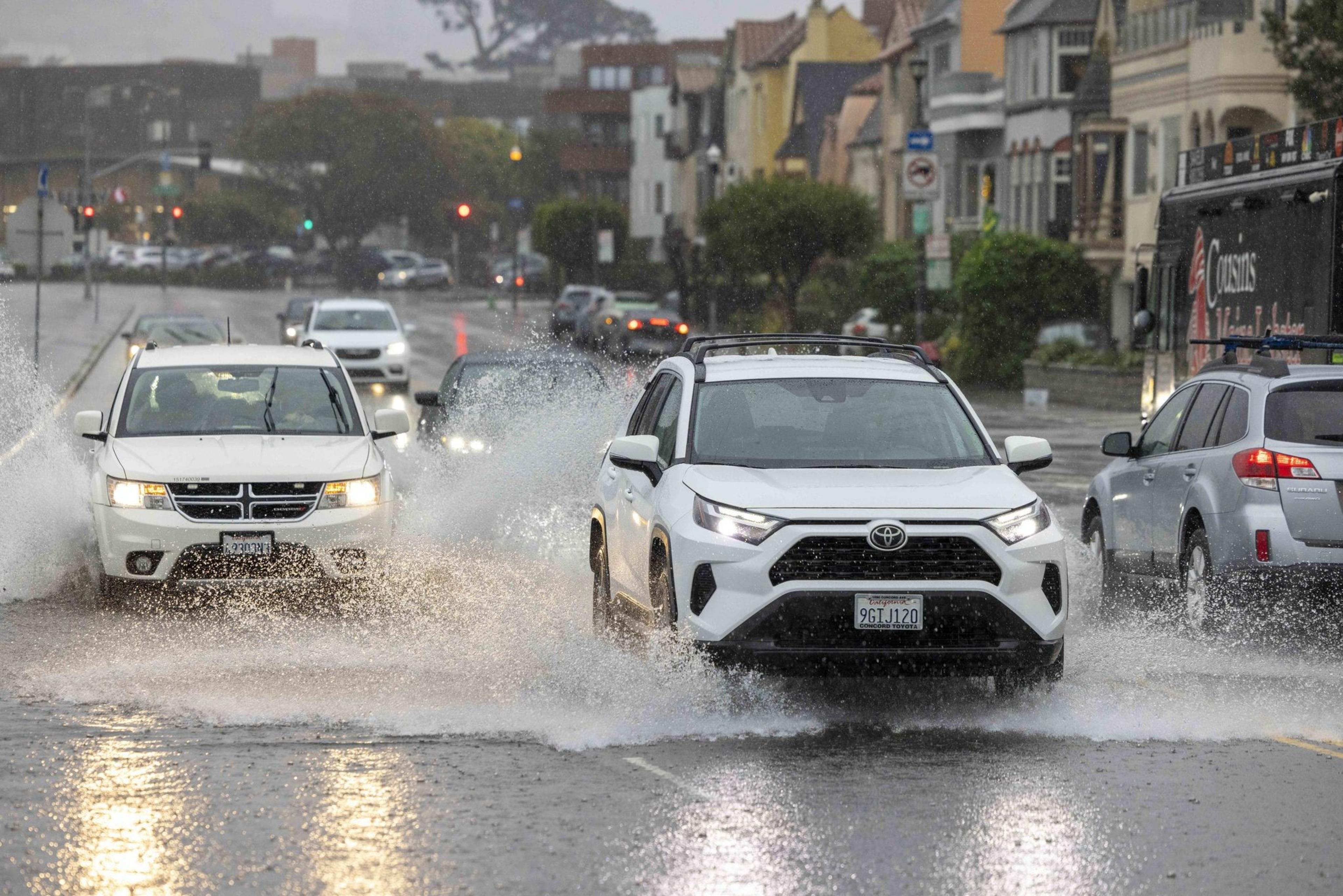 Cars drive through heavy rain and water splashes on a flooded street in a city, with buildings and traffic lights in the background.
