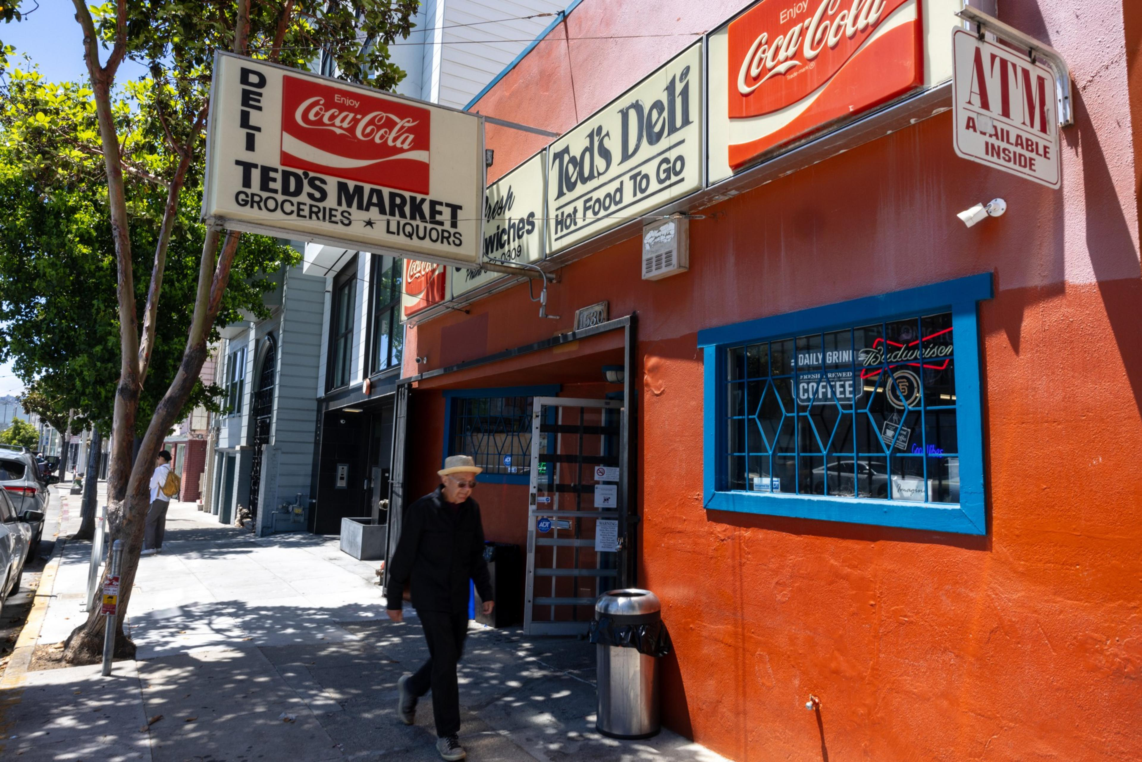 Ted’s Market deli with red and white Coca-Cola signs, an ATM inside, and a man walking on the sidewalk in front of the orange building on a sunny day.