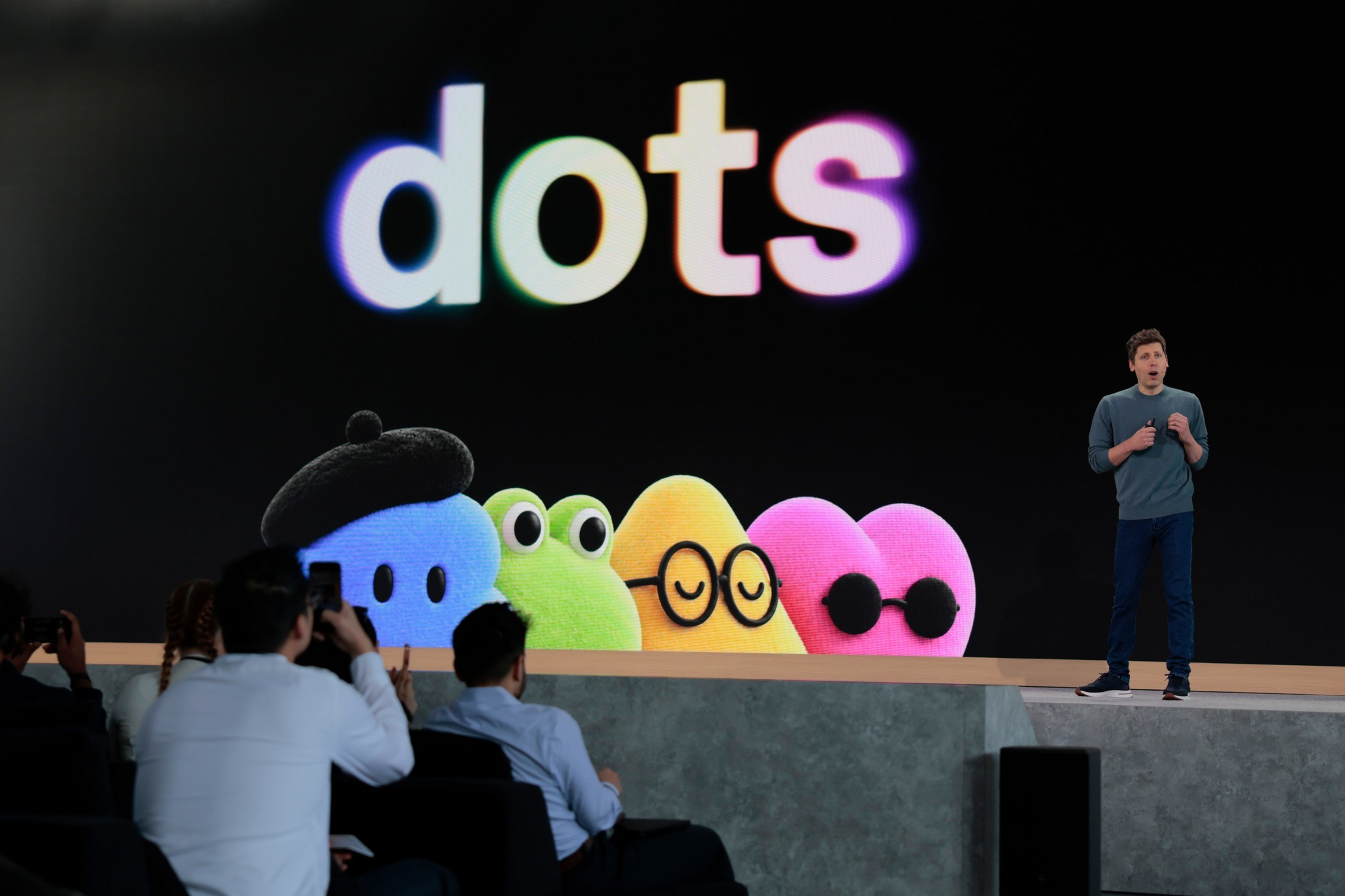 A presenter stands beside a large screen displaying the word "dots" in colorful letters and five colorful cartoon dots with various accessories and expressions.
