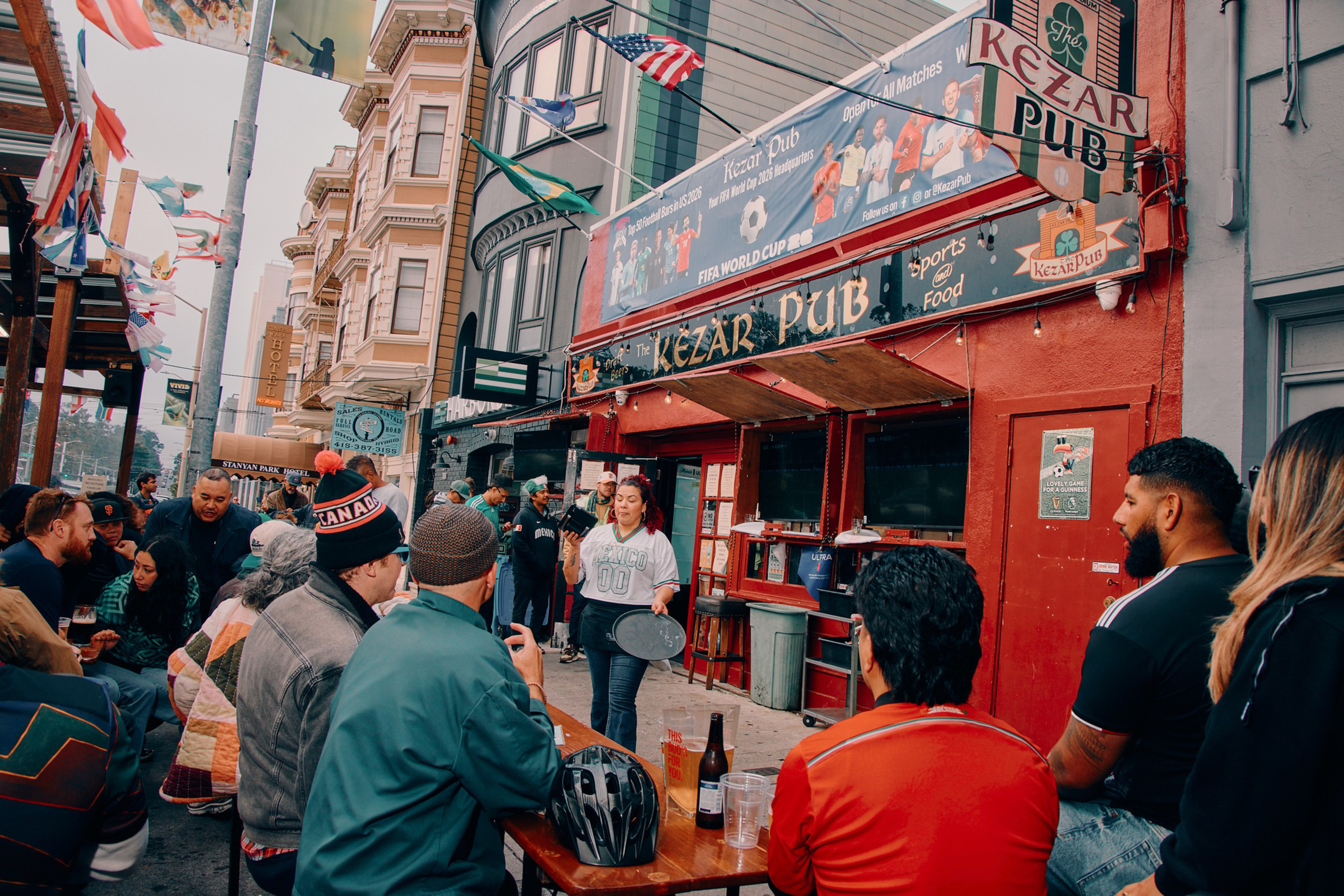 People gather outside a red building named Kezar Pub, sitting at tables and socializing, with flags hanging above the street in a lively urban setting.