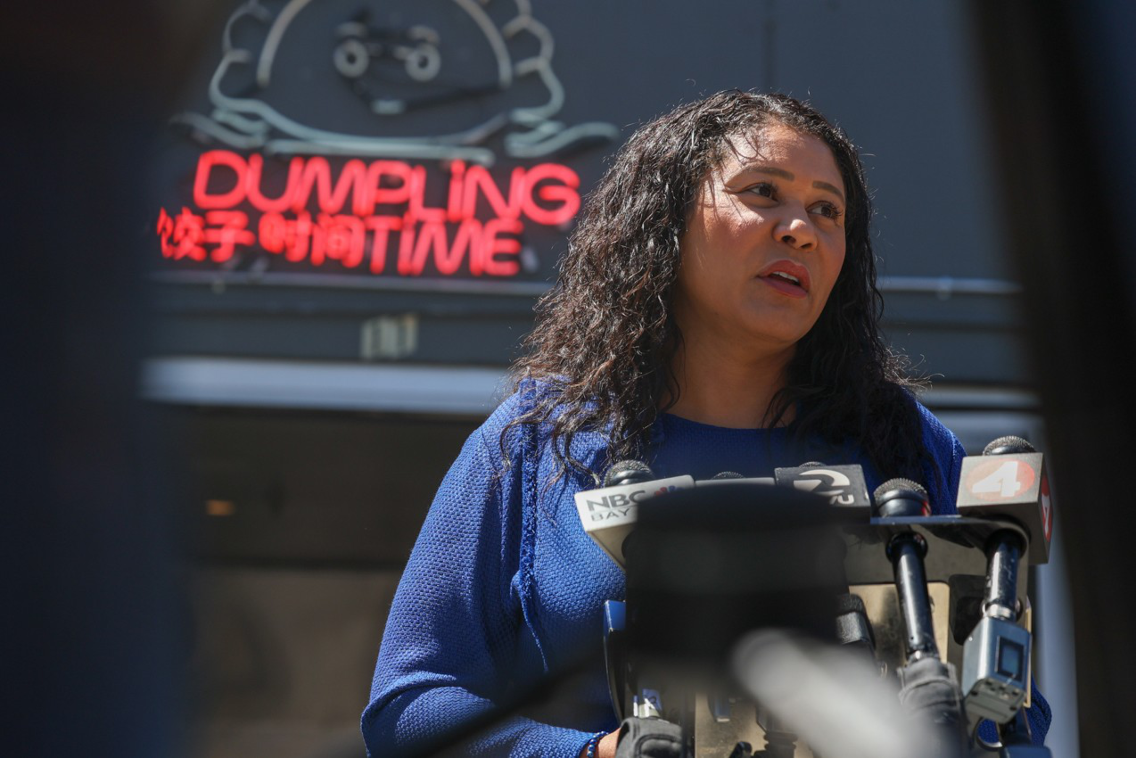 A woman with curly hair in a blue top speaks at a podium with multiple microphones in front of a neon sign reading "DUMPLING TIME."
