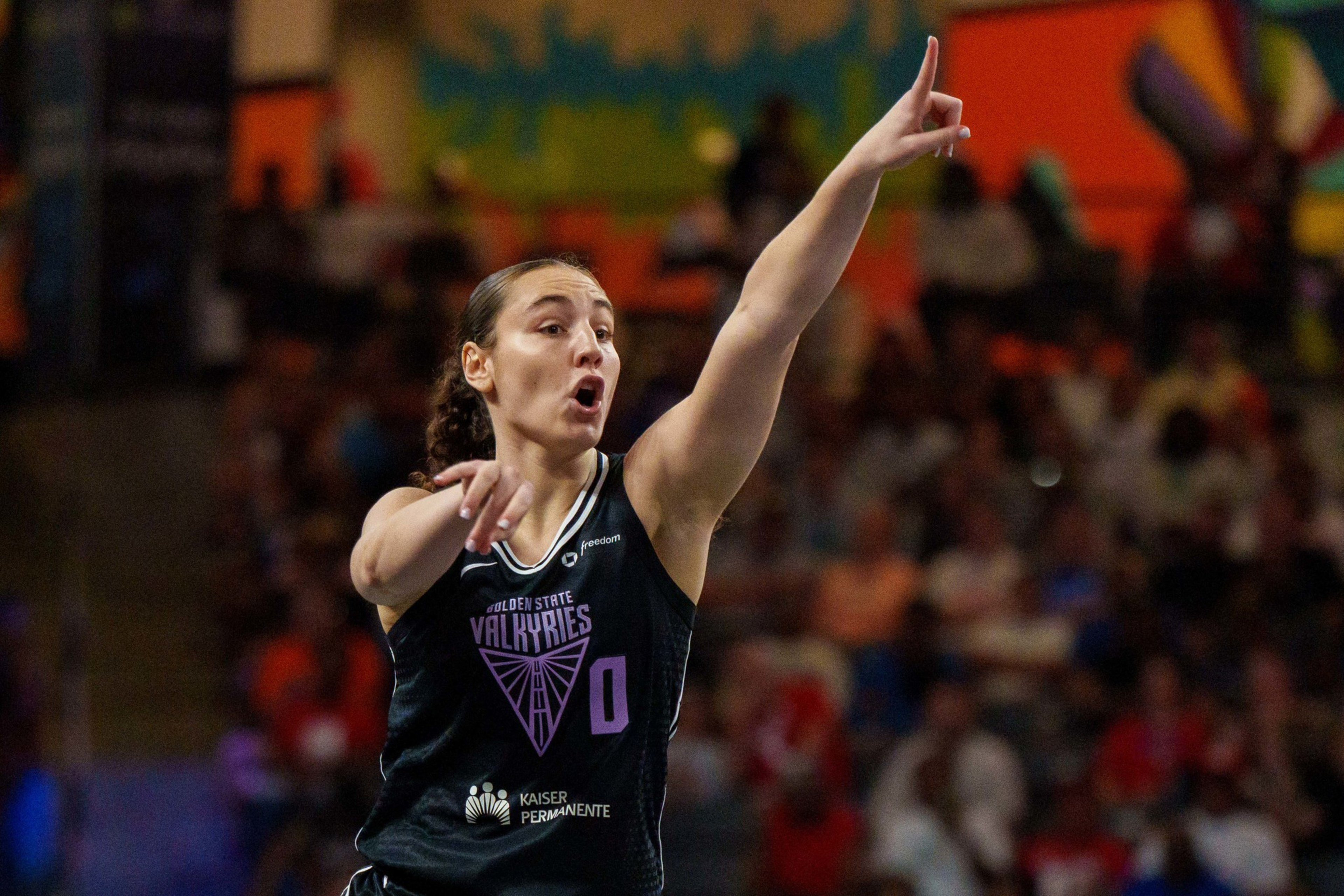 A basketball player in a black Golden State Valkyries jersey with number 0 points and directs with her right arm raised.