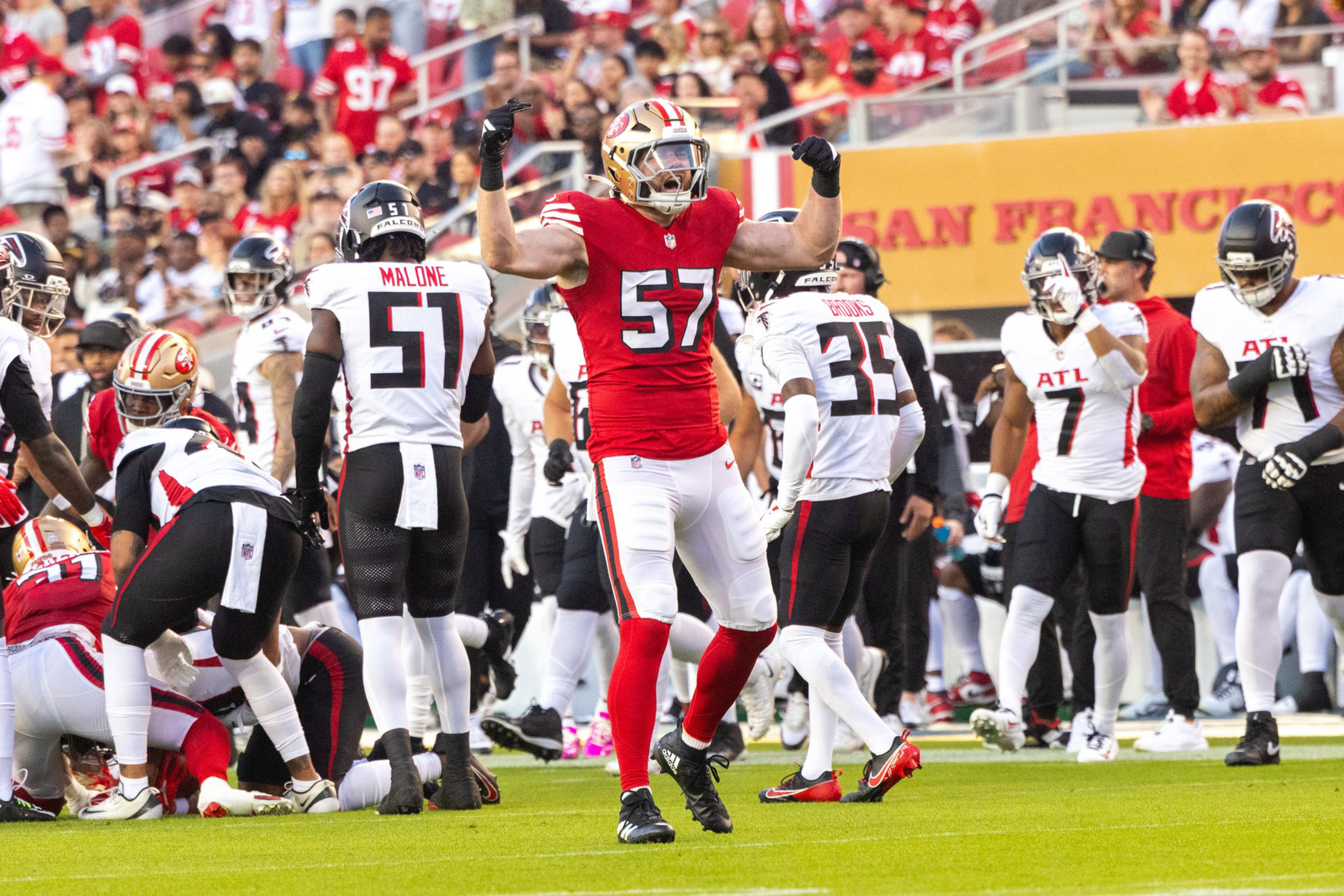 A football player in a red jersey with number 57 is celebrating with arms raised while players in white and black jerseys stand nearby on the field.
