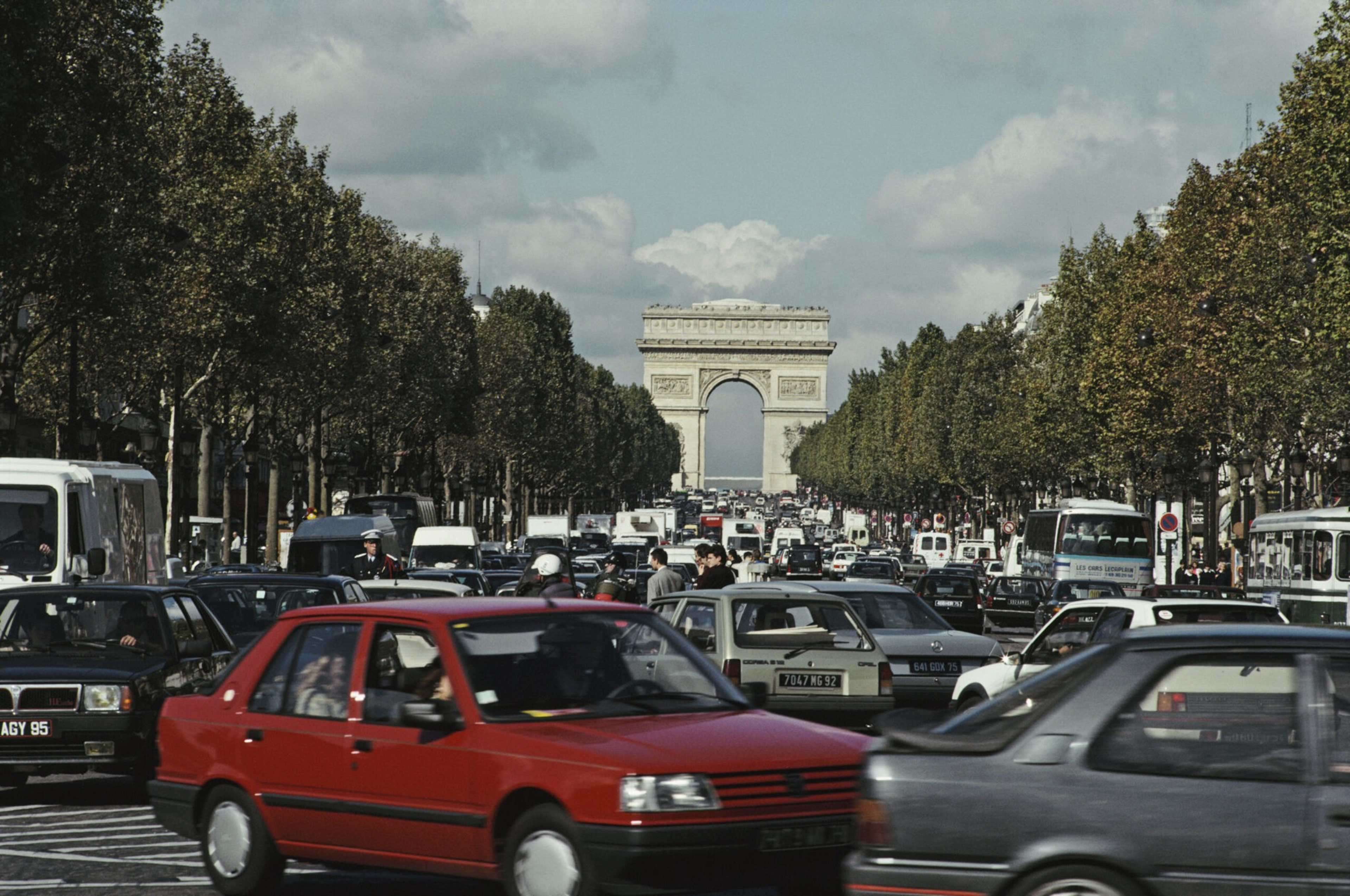Heavy traffic fills a broad tree-lined avenue leading to the Arc de Triomphe under a cloudy sky.
