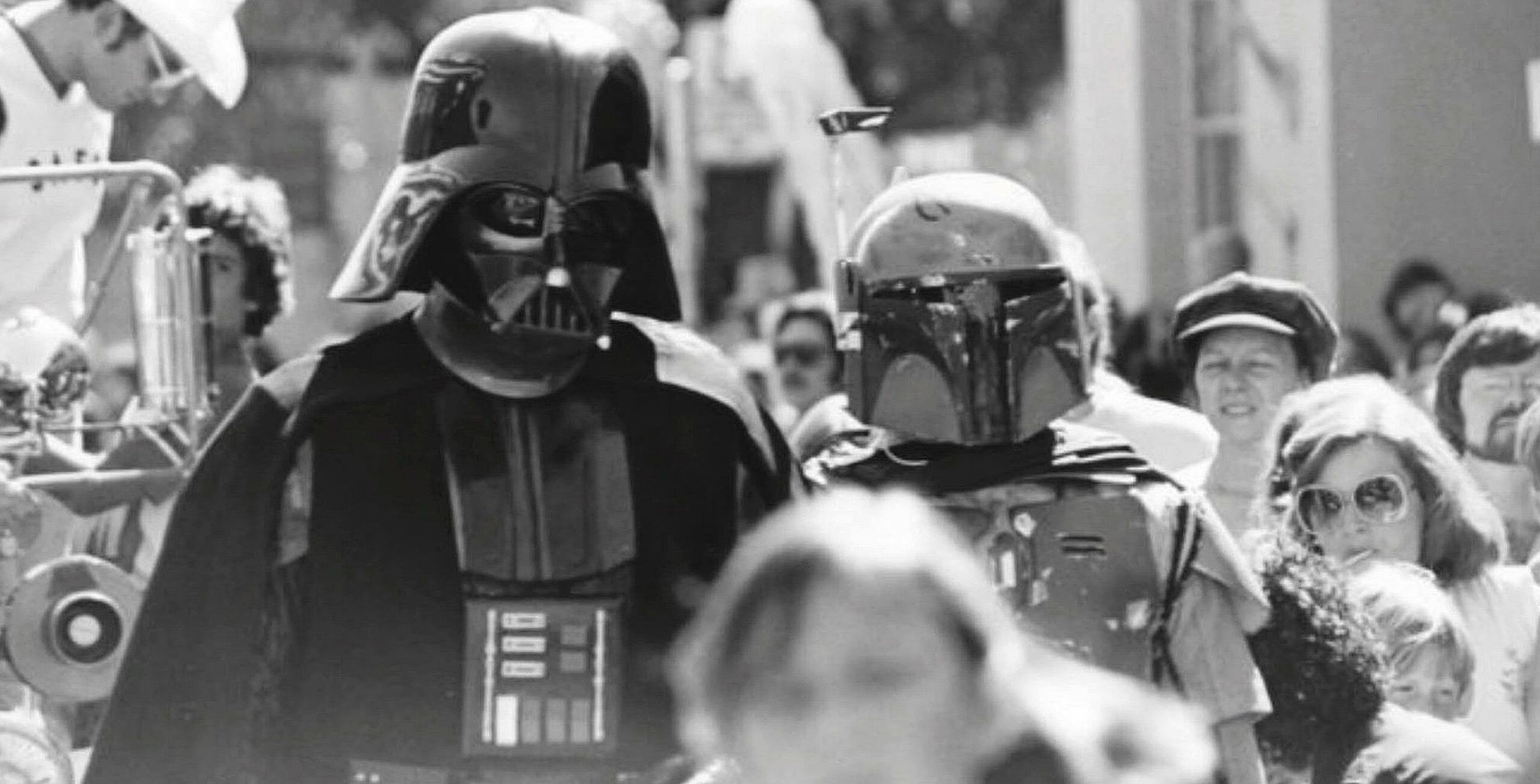 Two people dressed as Darth Vader and Boba Fett from Star Wars walk among a crowd of casually dressed onlookers outdoors.