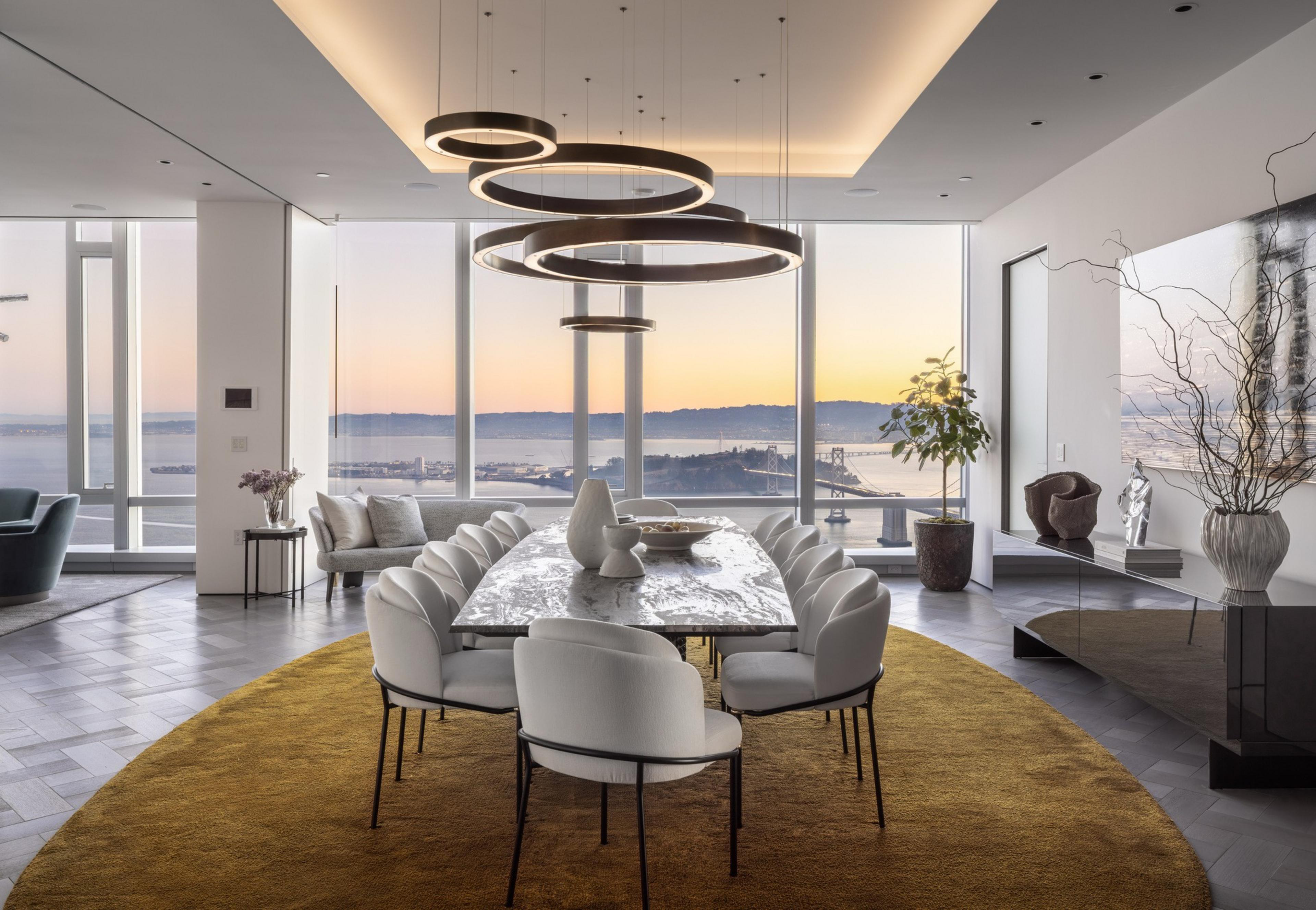 A modern dining room features a long marble table surrounded by white chairs on a mustard rug, with large windows offering a sunset view of a bridge and water.
