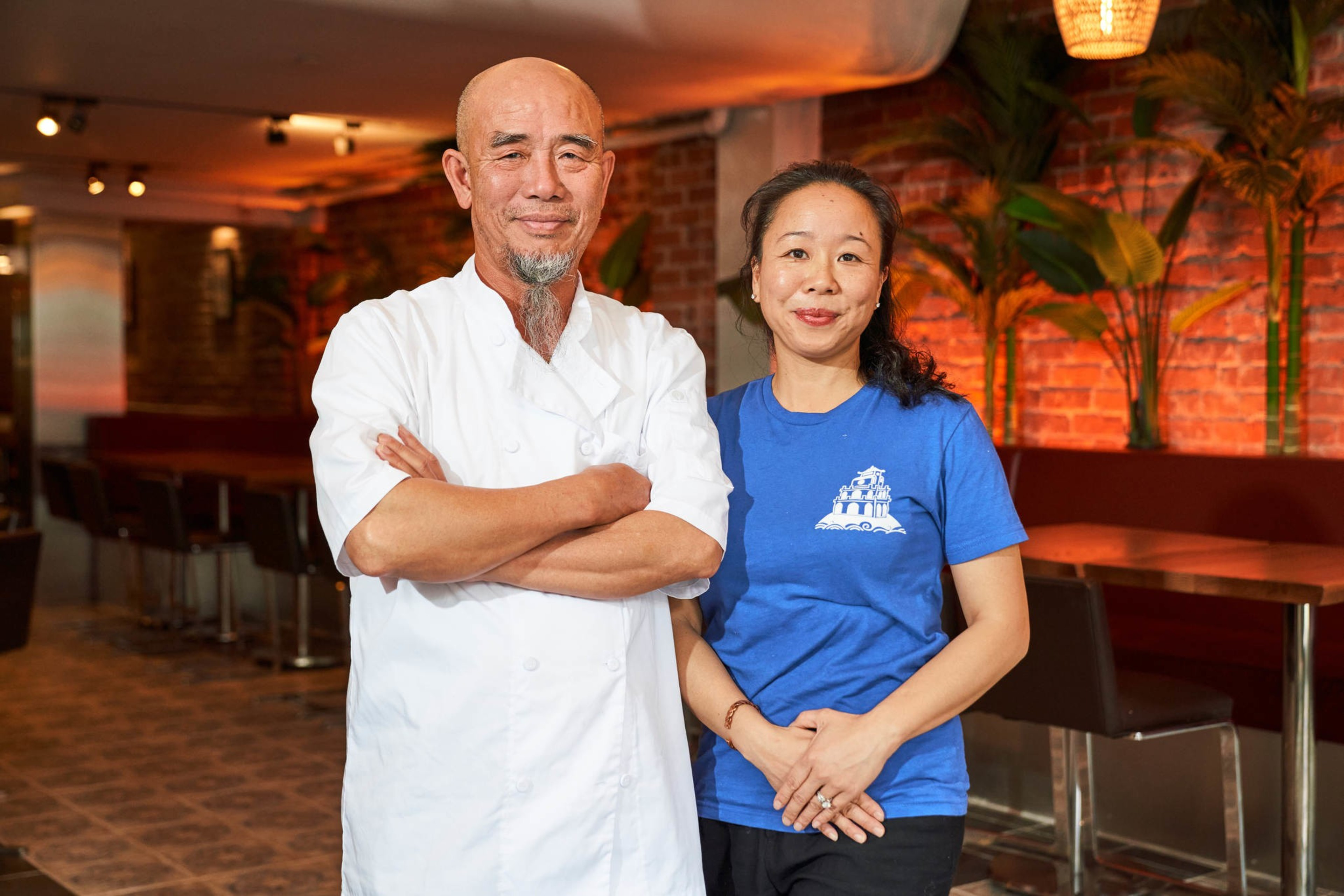 A man in a chef's jacket and a woman in a blue shirt stand smiling in a restaurant with red brick walls and plants. The setting is warmly lit.