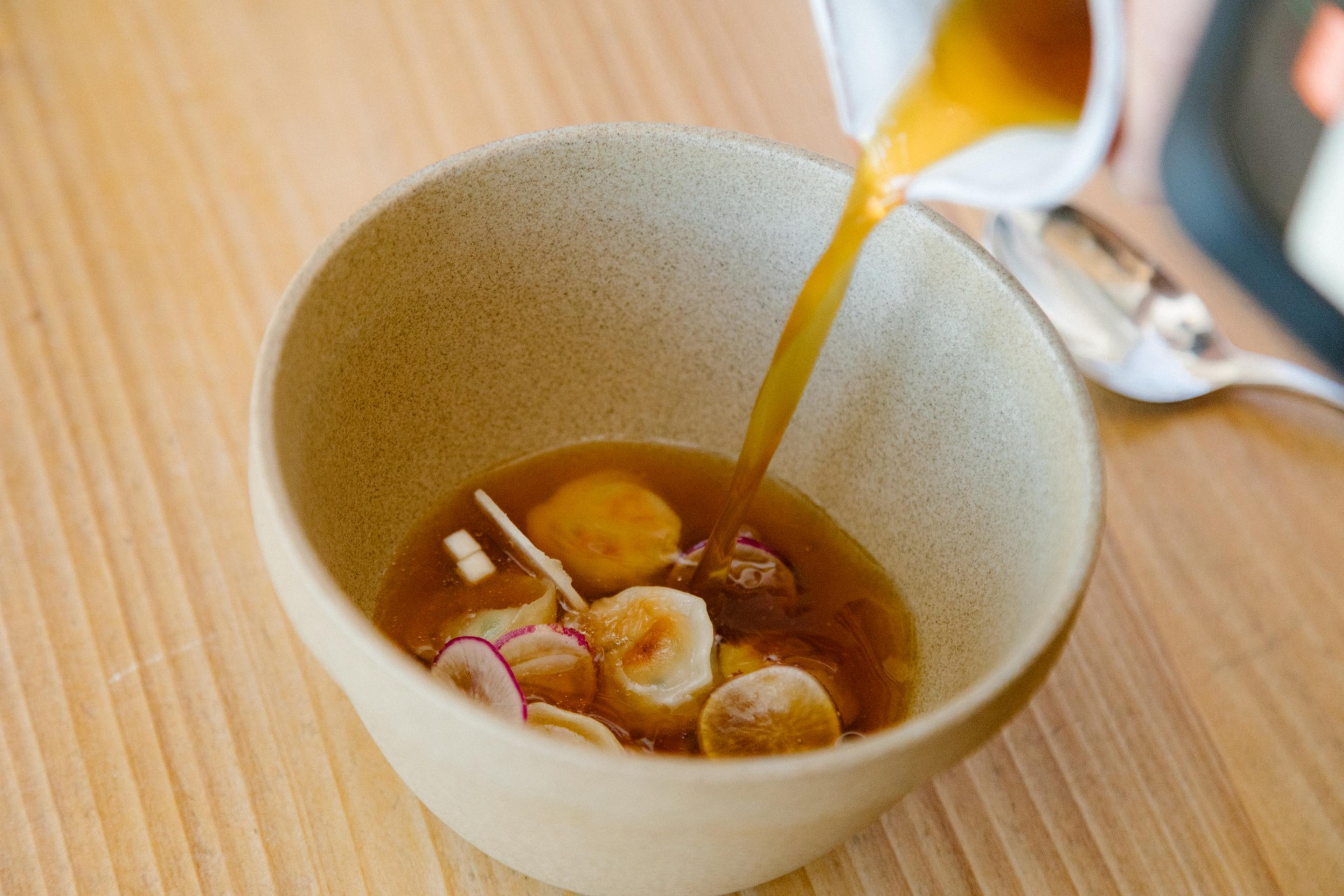 A light brown broth is being poured into a bowl containing sliced bananas and thinly sliced red onions on a wooden surface.