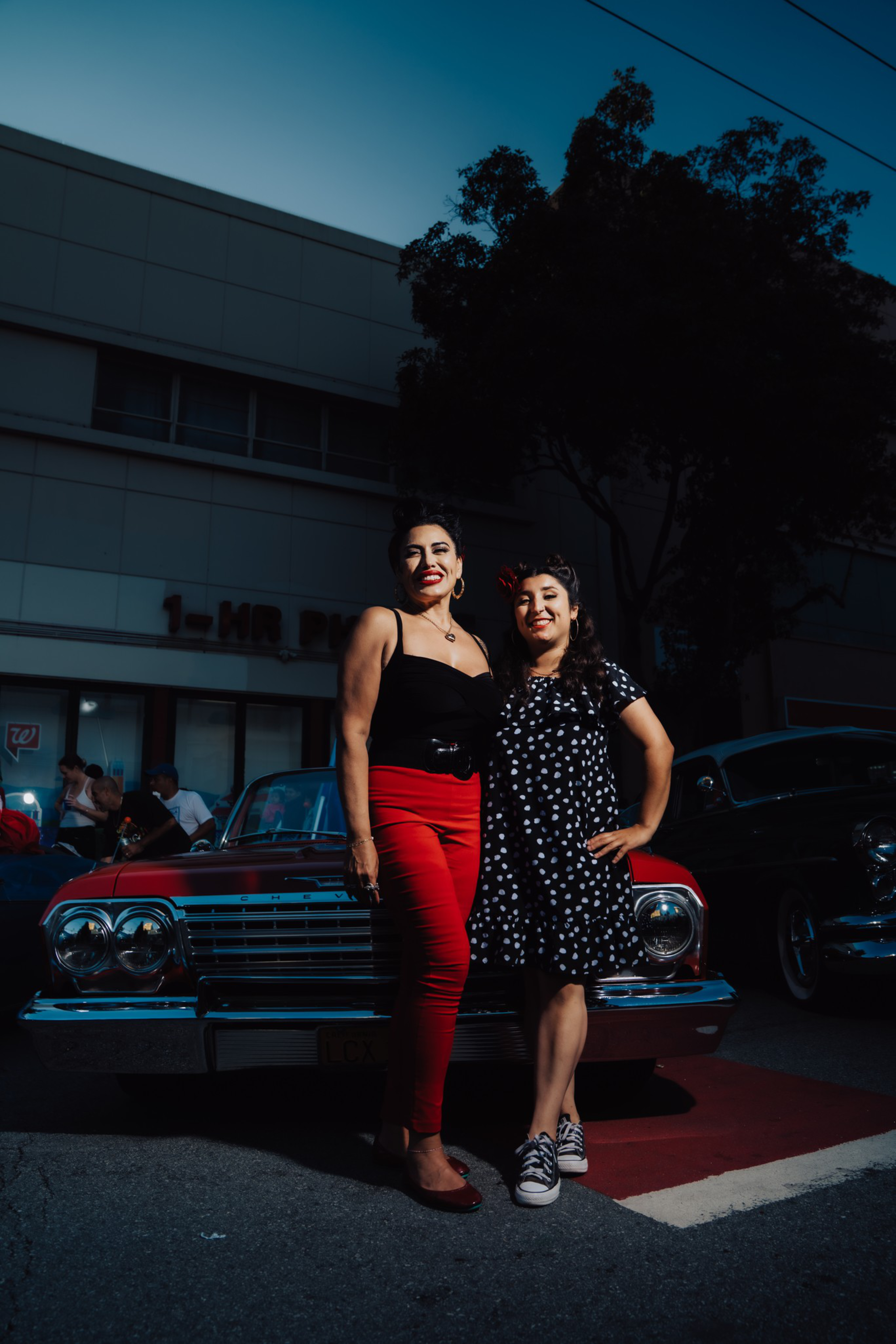 Two women smile and pose in front of a classic red car parked on the street at dusk, with a building and trees in the background.