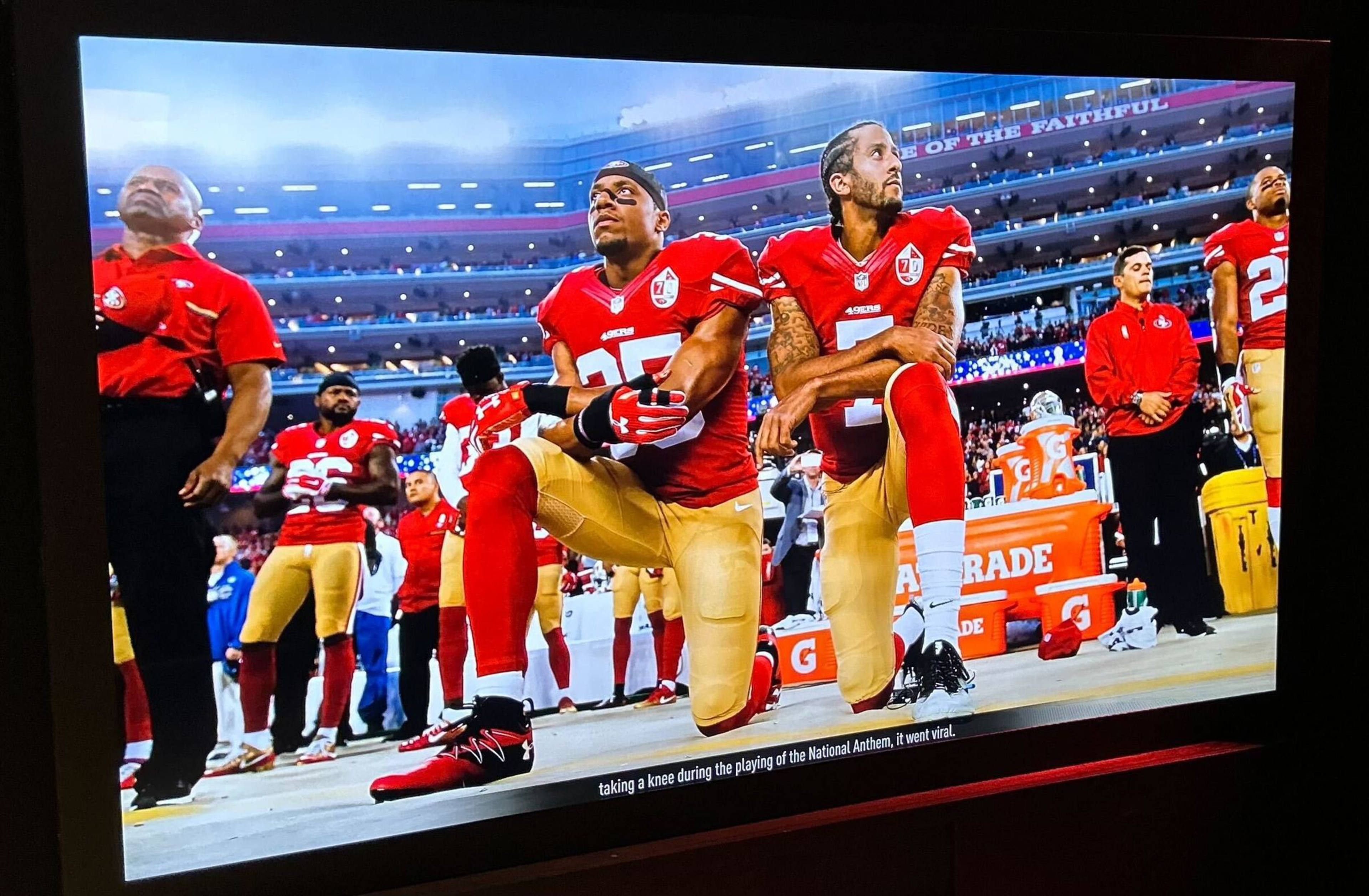 Two football players in red and gold uniforms kneel side by side on the field, while others stand behind them near the sidelines.
