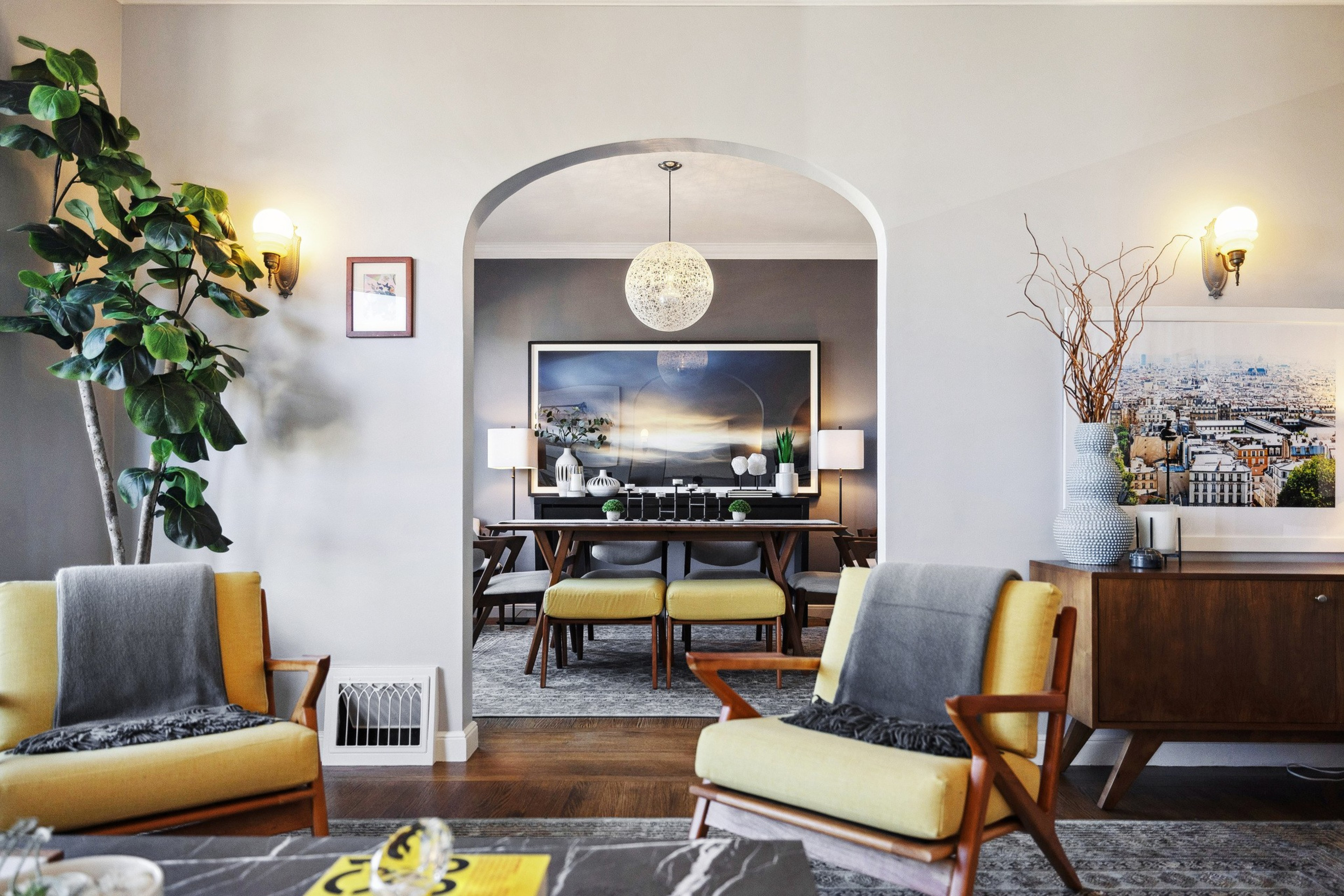 A cozy living room with yellow chairs, a large indoor plant, wall sconces, and wooden furniture, extending into a dining area through an arched doorway.