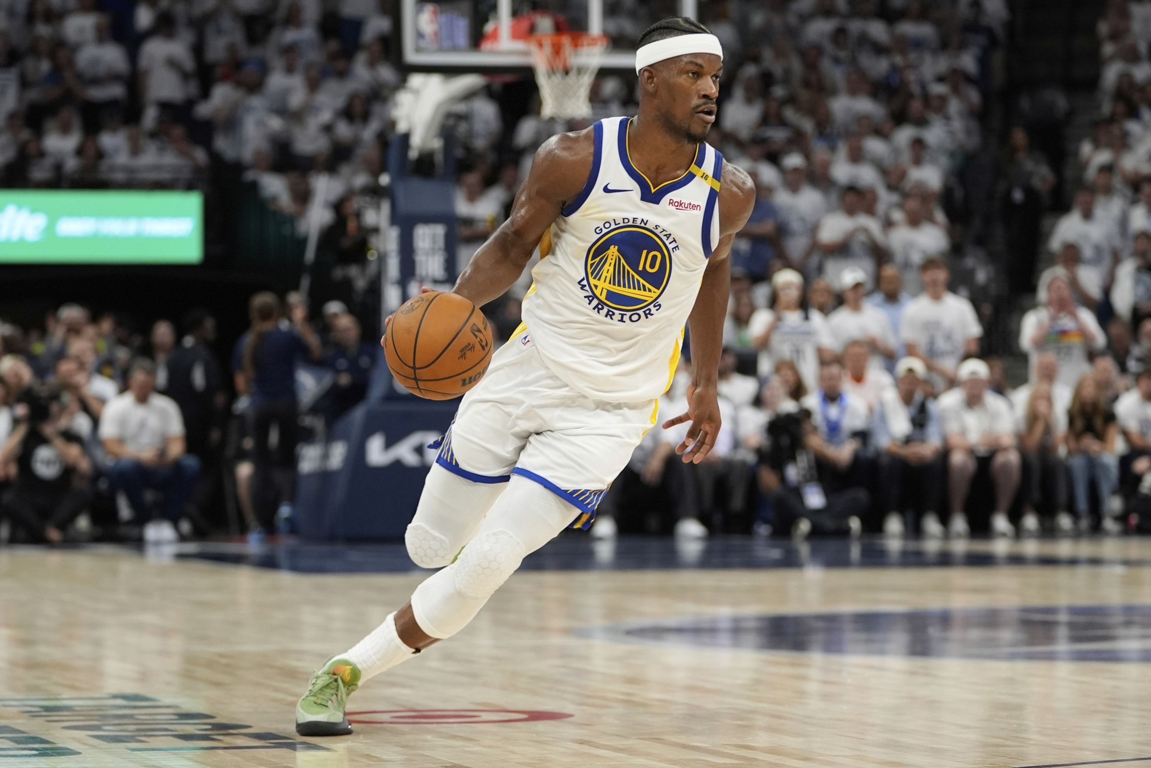 A basketball player in a Warriors jersey, number 10, is dribbling the ball on the court. The crowd in the background watches intently.