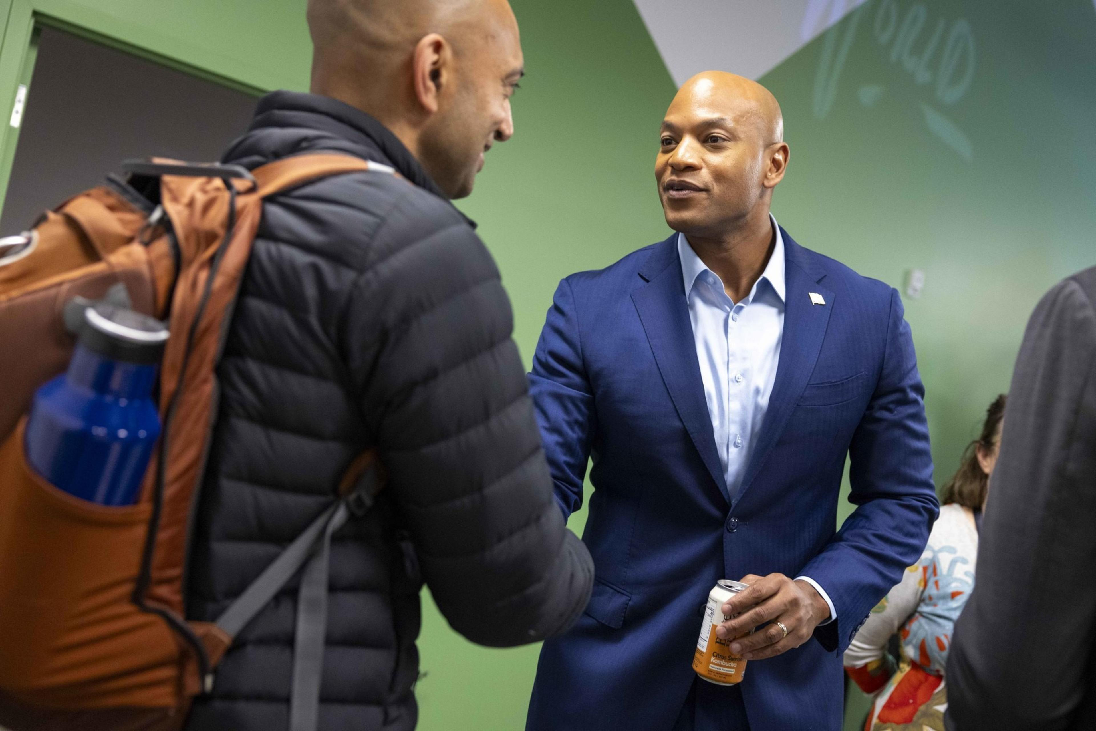 A man in a blue suit shakes hands with another man wearing a backpack and holding a blue water bottle, against a green wall.