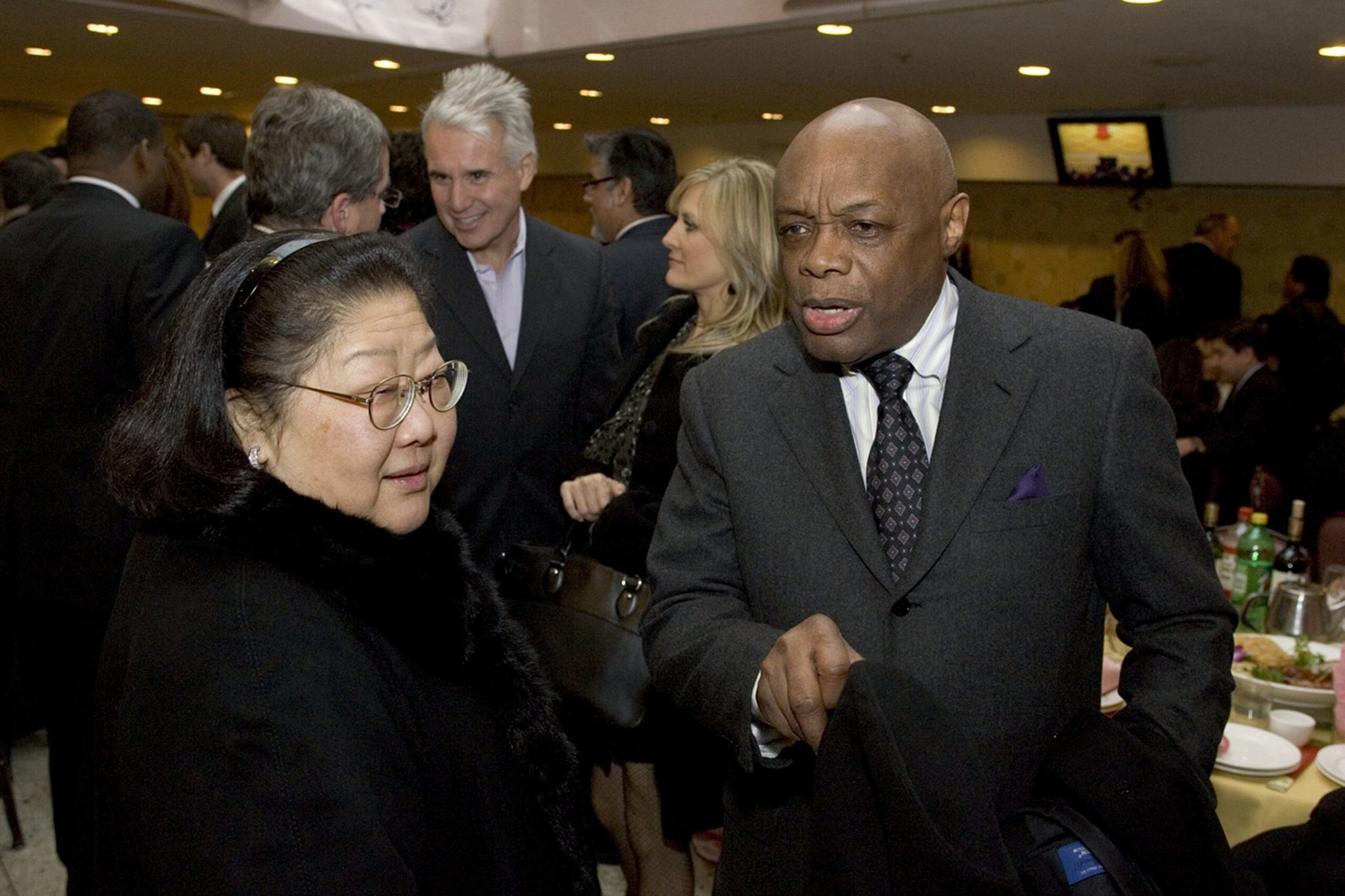A man in a dark suit speaks while holding a coat; a woman with glasses and a headband listens, with other people mingling in the background.