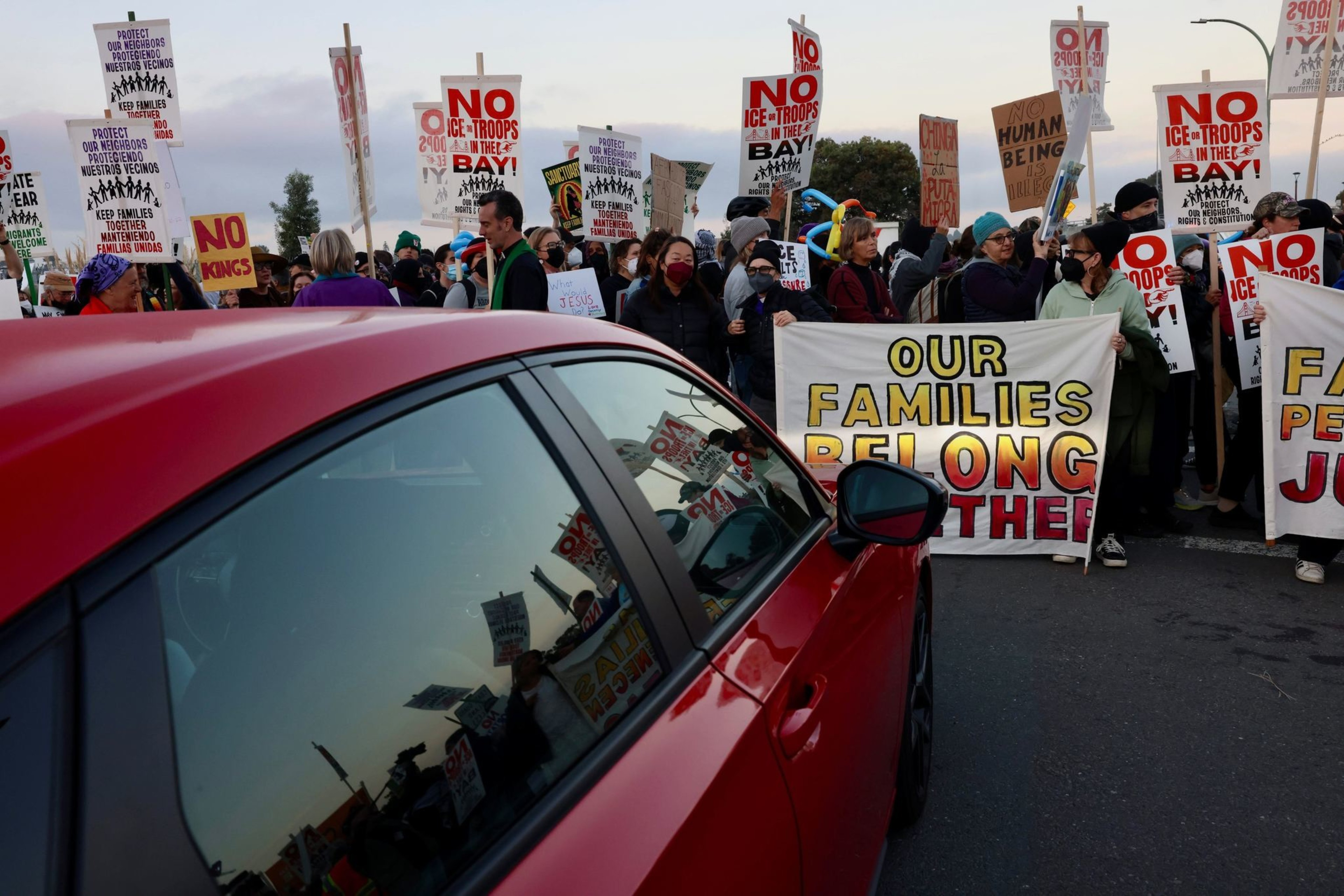 A crowd of protesters holds signs with messages like “Our families belong together” and “No ICE troops in the bay,” standing in front of a red car.
