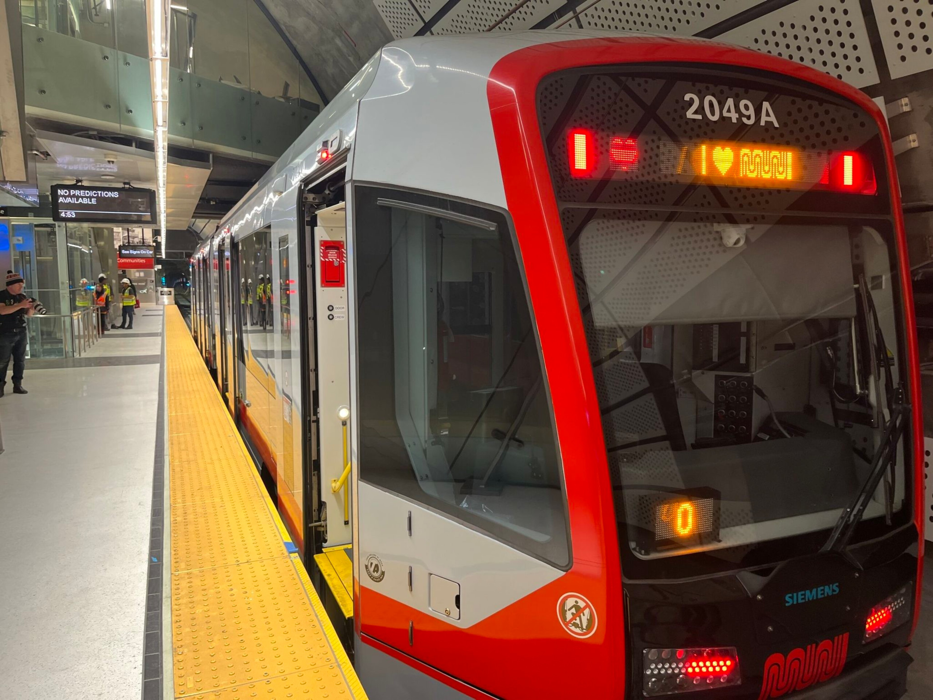 A modern red and grey Muni train with open doors is stationed at a clean underground platform with a yellow safety strip and few people nearby.