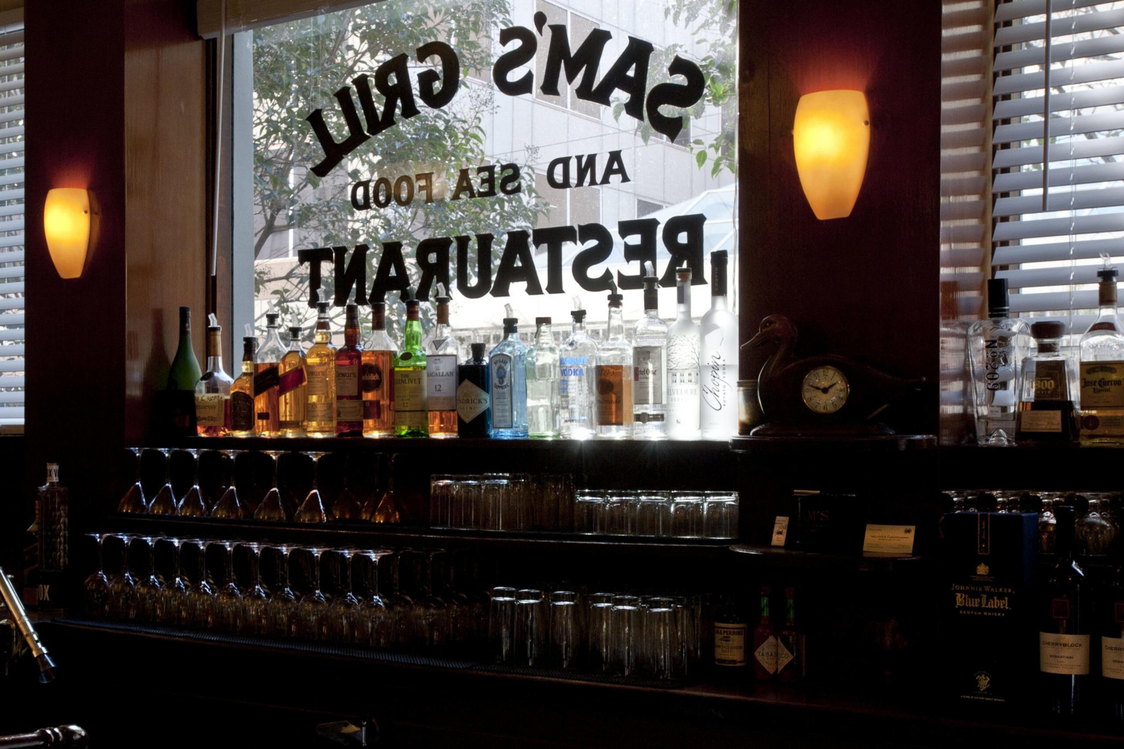 Shelves of liquor bottles, glasses, and a vintage clock are backlit by a window with "Sam's Grill and Seafood Restaurant" painted on it.