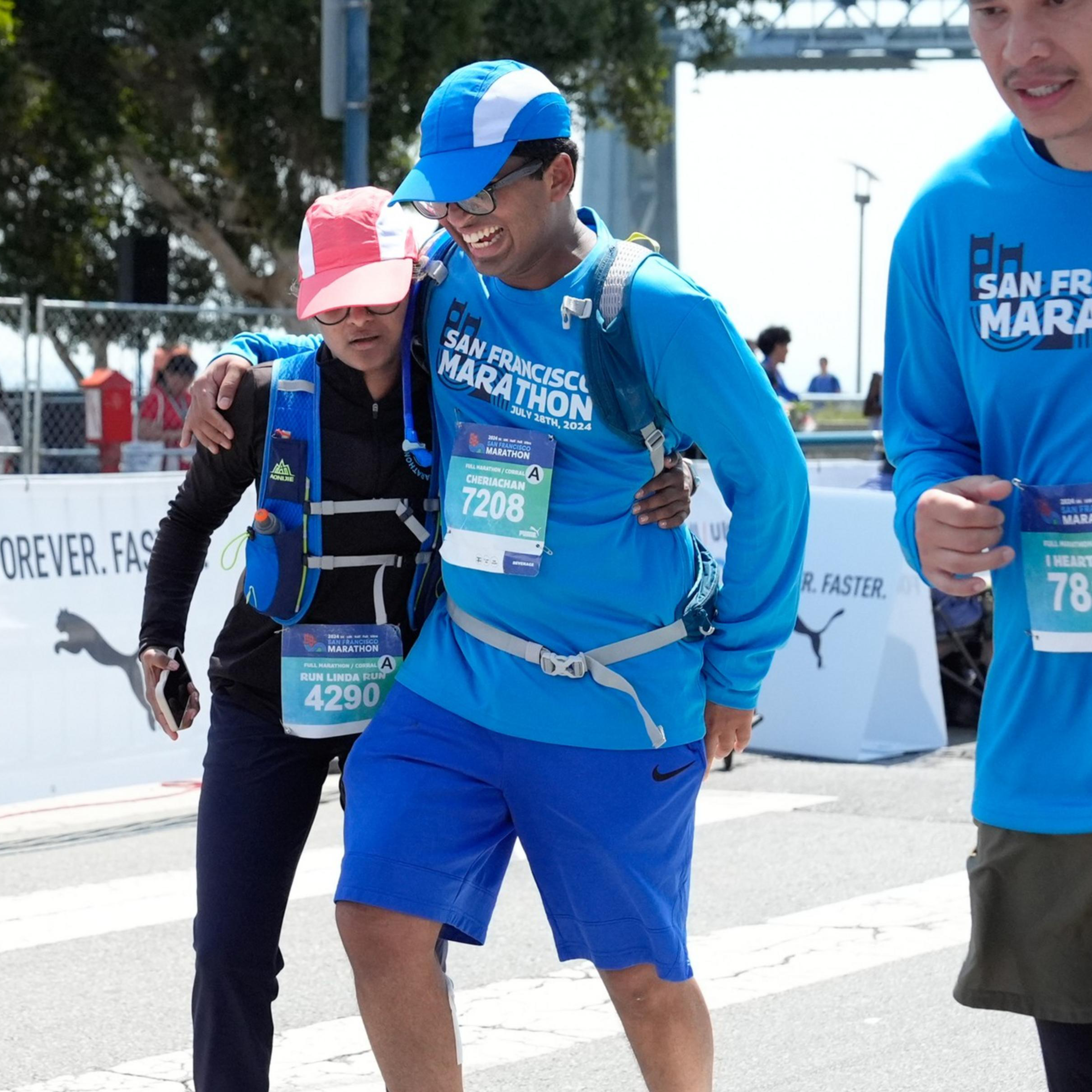 Three runners wearing blue “San Francisco Marathon” shirts cross the finish line together, two of them assisting each other while smiling.