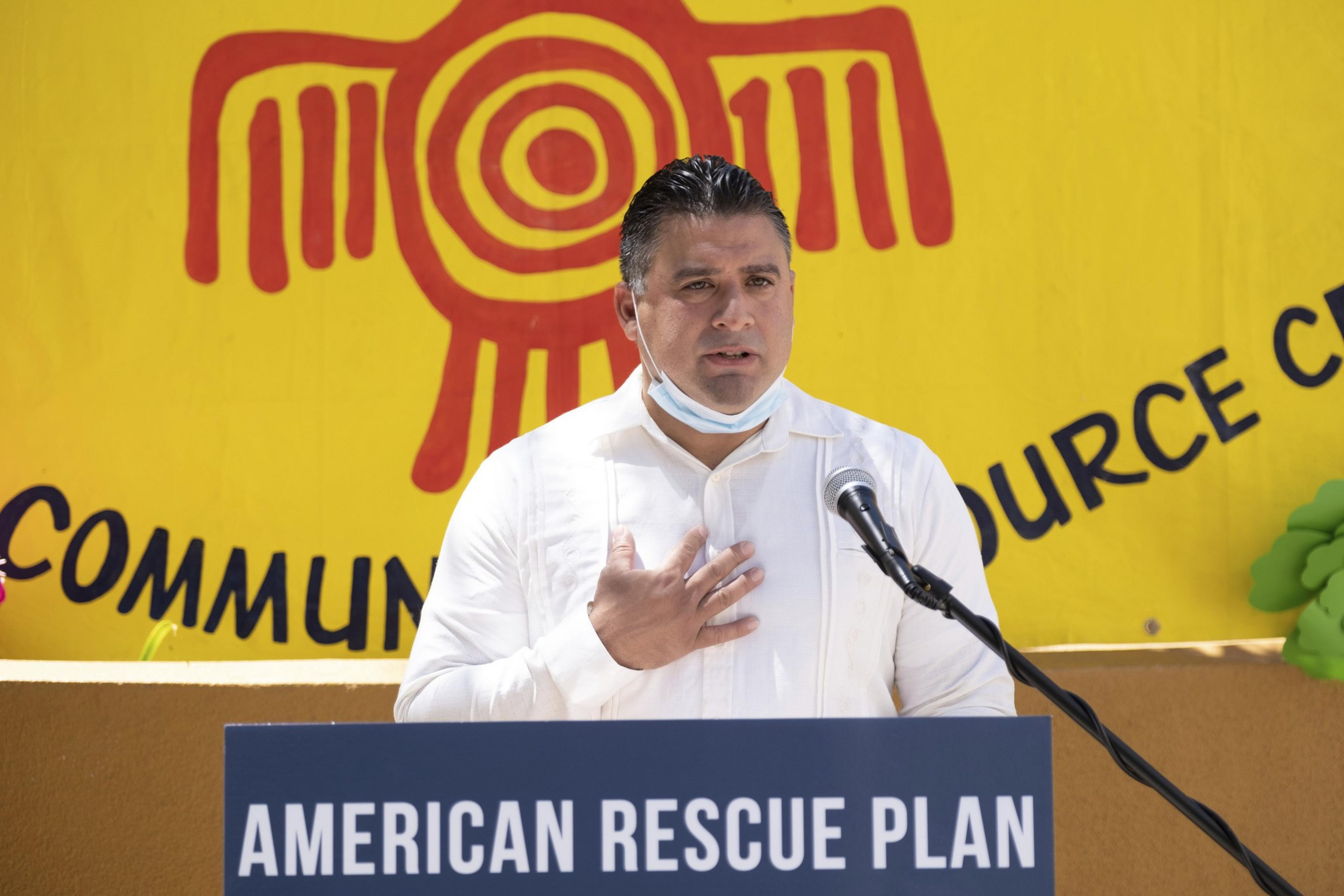 A man in a white shirt speaks at a podium labeled &quot;American Rescue Plan.&quot; He gestures with his hand on his chest, standing before a bright yellow backdrop.