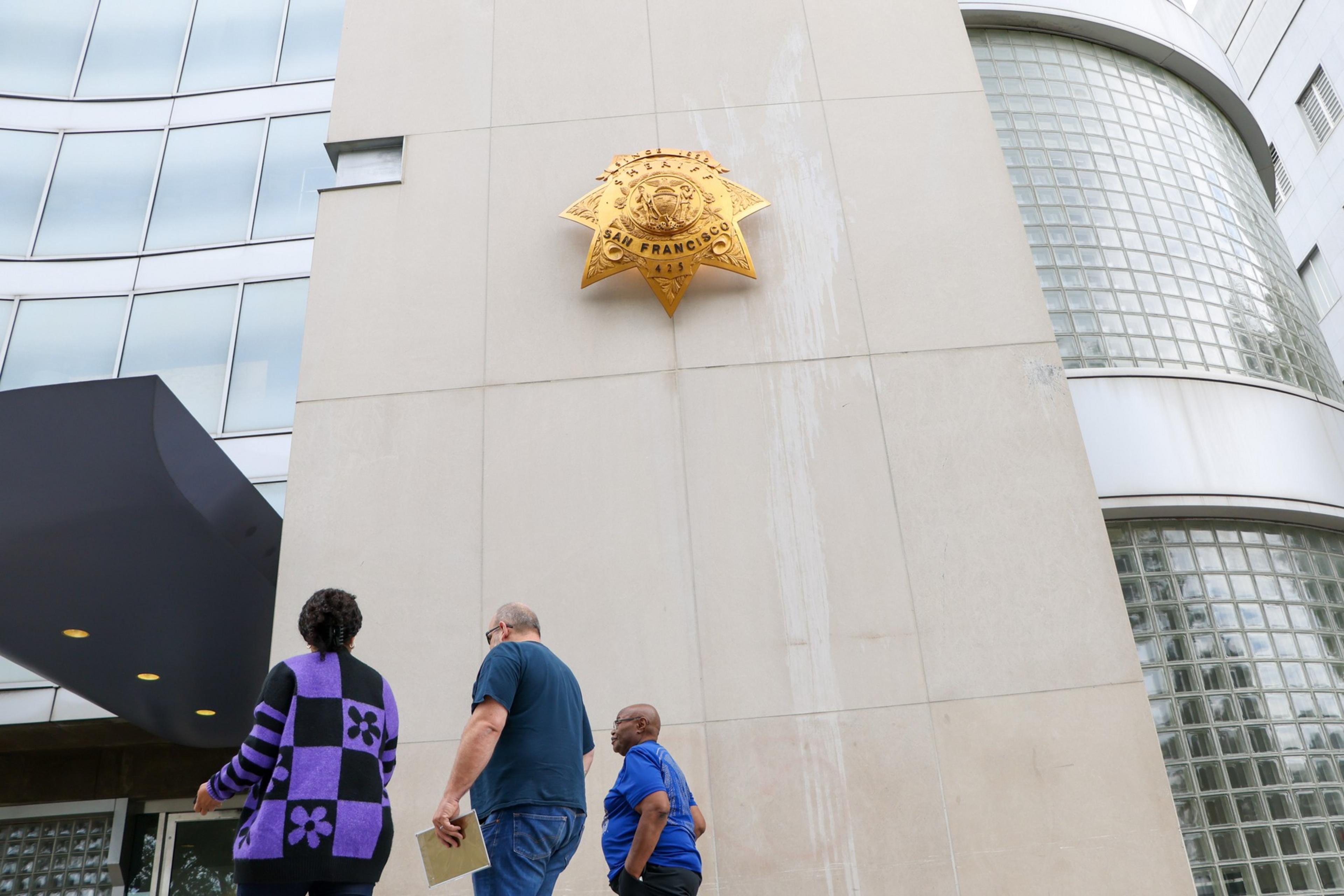 Three people walk past a building with a large badge emblem stating &quot;San Francisco Police&quot;.