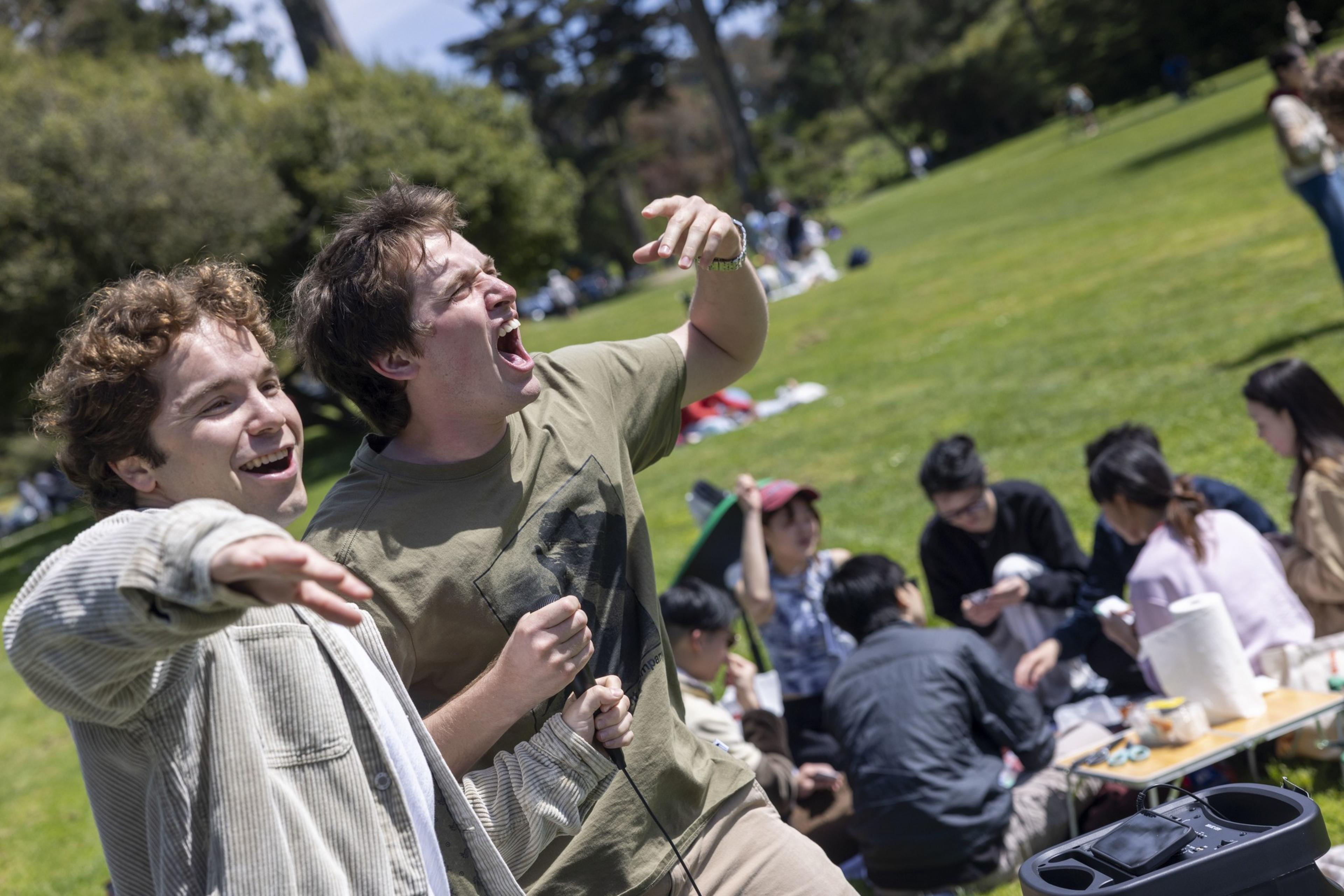 Two men joyfully sing with a microphone in a park, surrounded by people sitting on the grass with picnic items. The scene is lively and sunny.