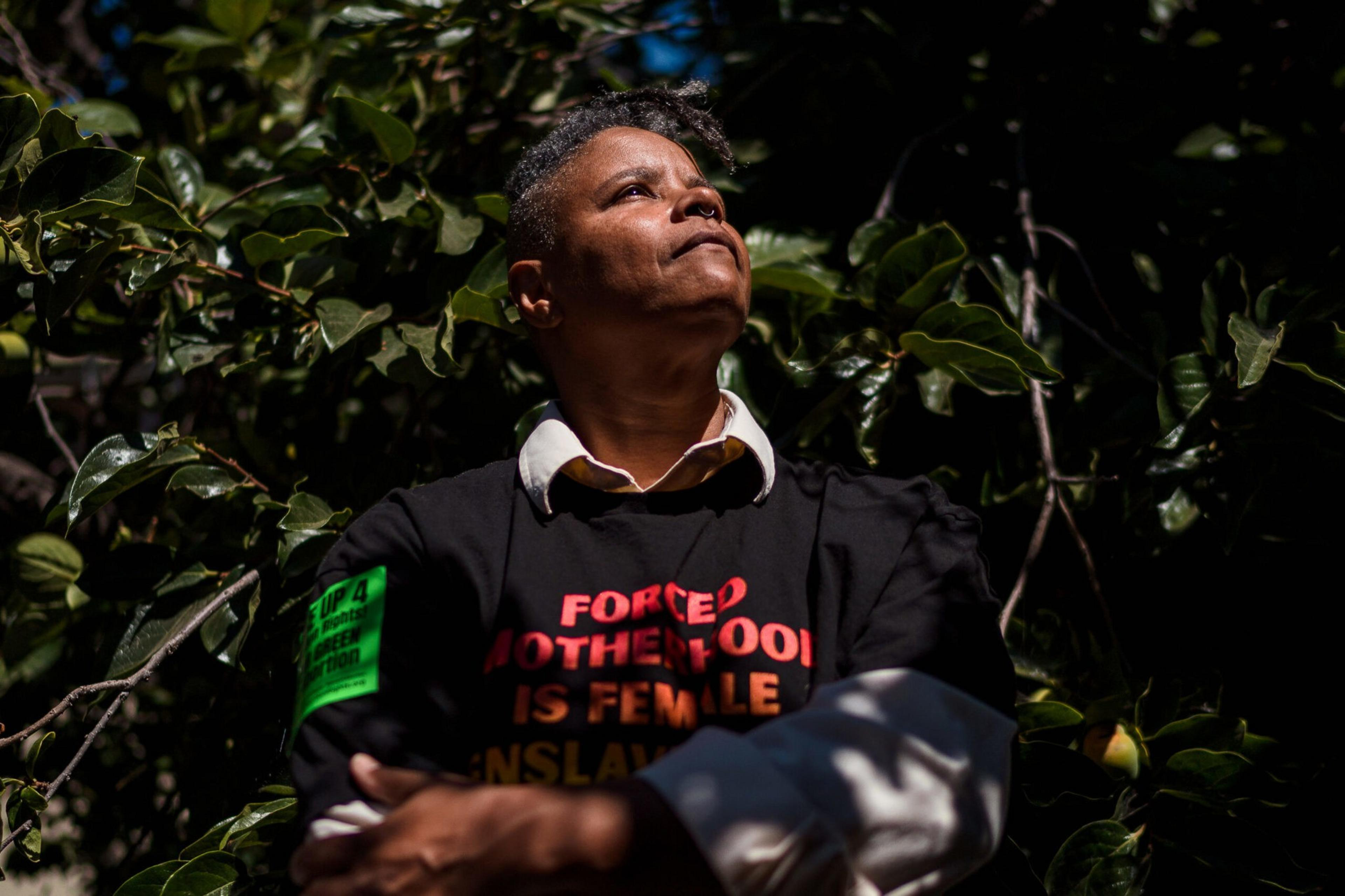 A person with short hair looks upward, wearing a black shirt with red and orange text and a green patch on the sleeve, surrounded by dark green foliage.