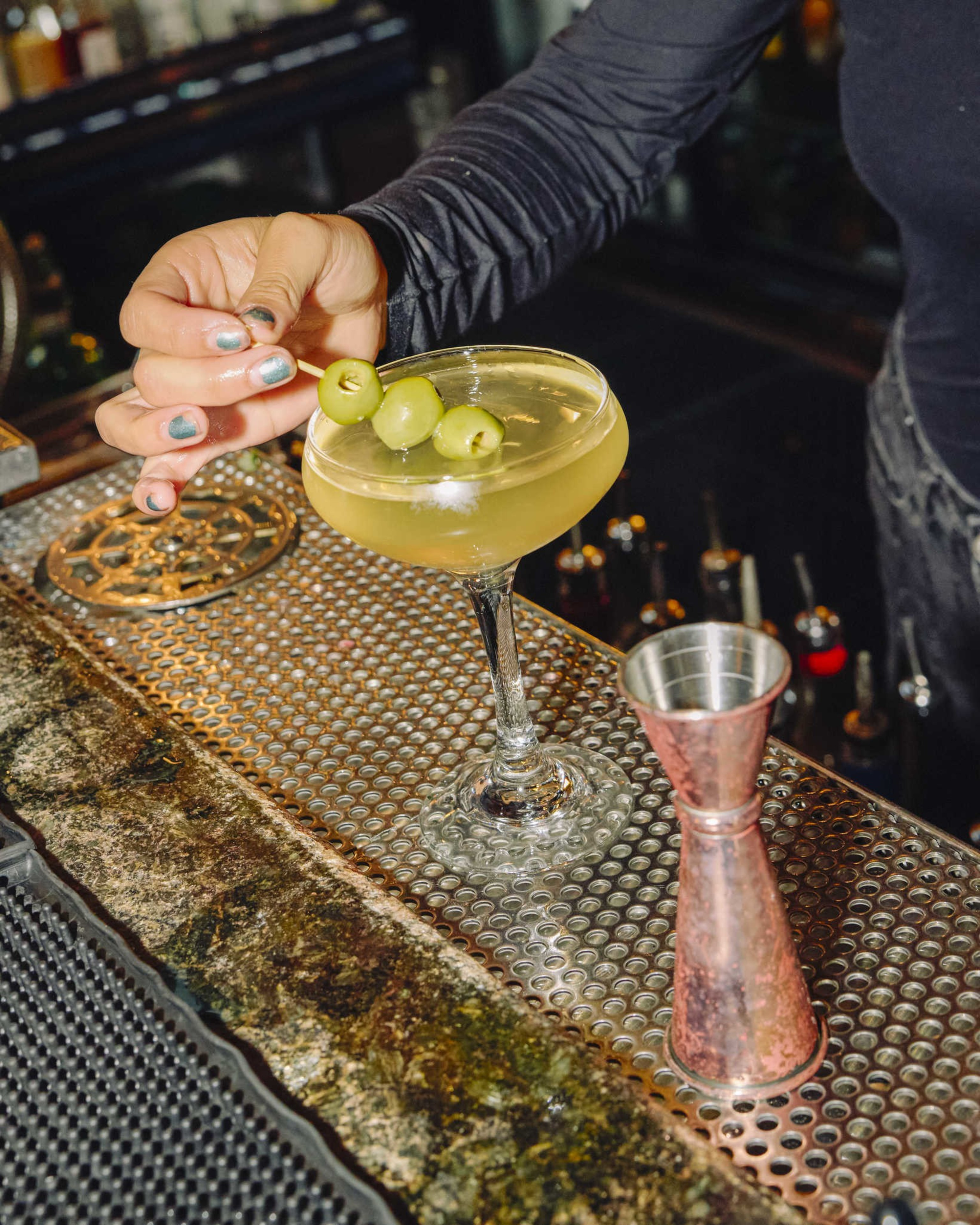 A hand with painted nails is garnishing a cocktail glass filled with a light green drink using a skewer of three green olives on a bar counter.