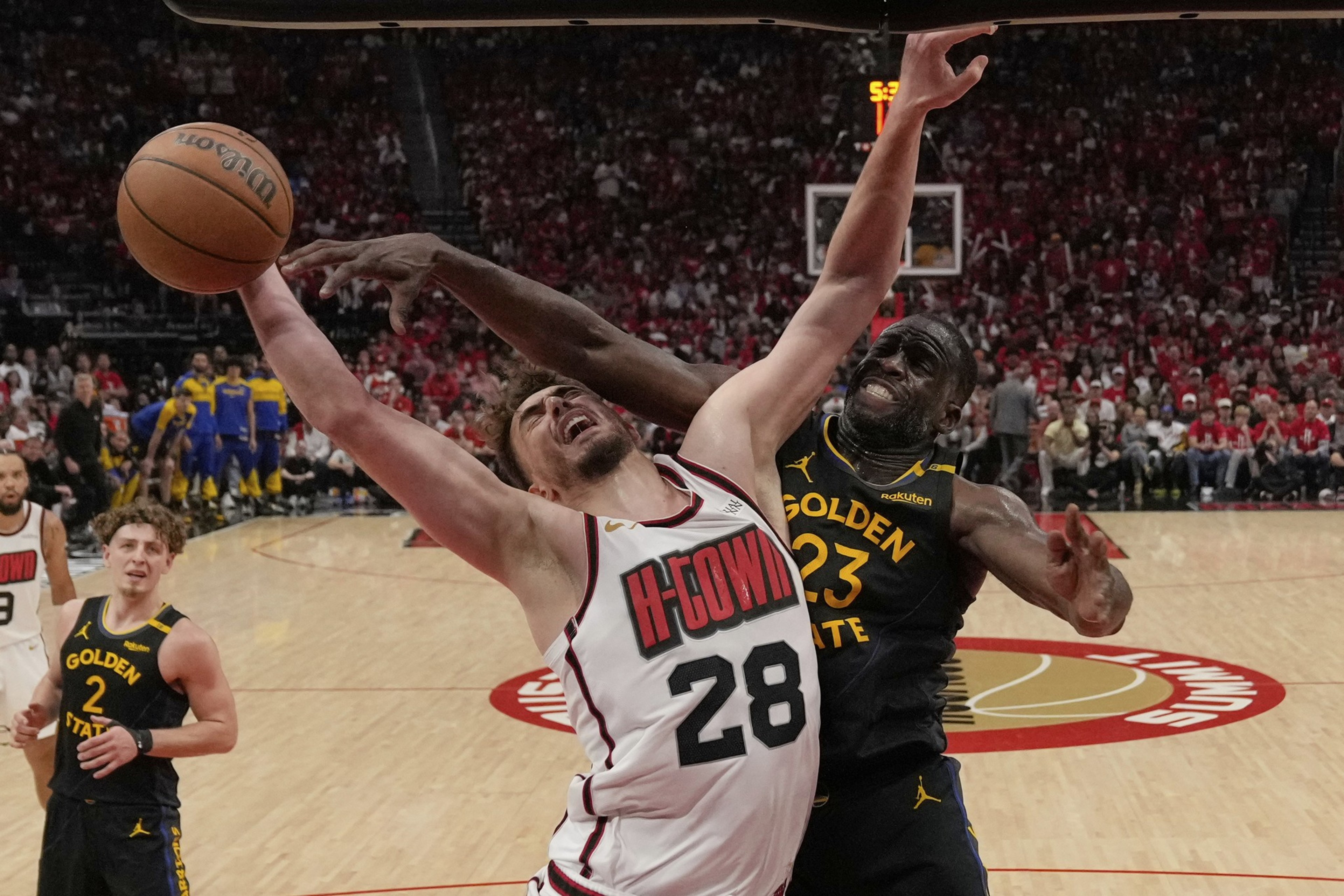 A basketball player in a white jersey is attempting a shot while being blocked by another player in a black jersey. The crowd is blurred in the background.