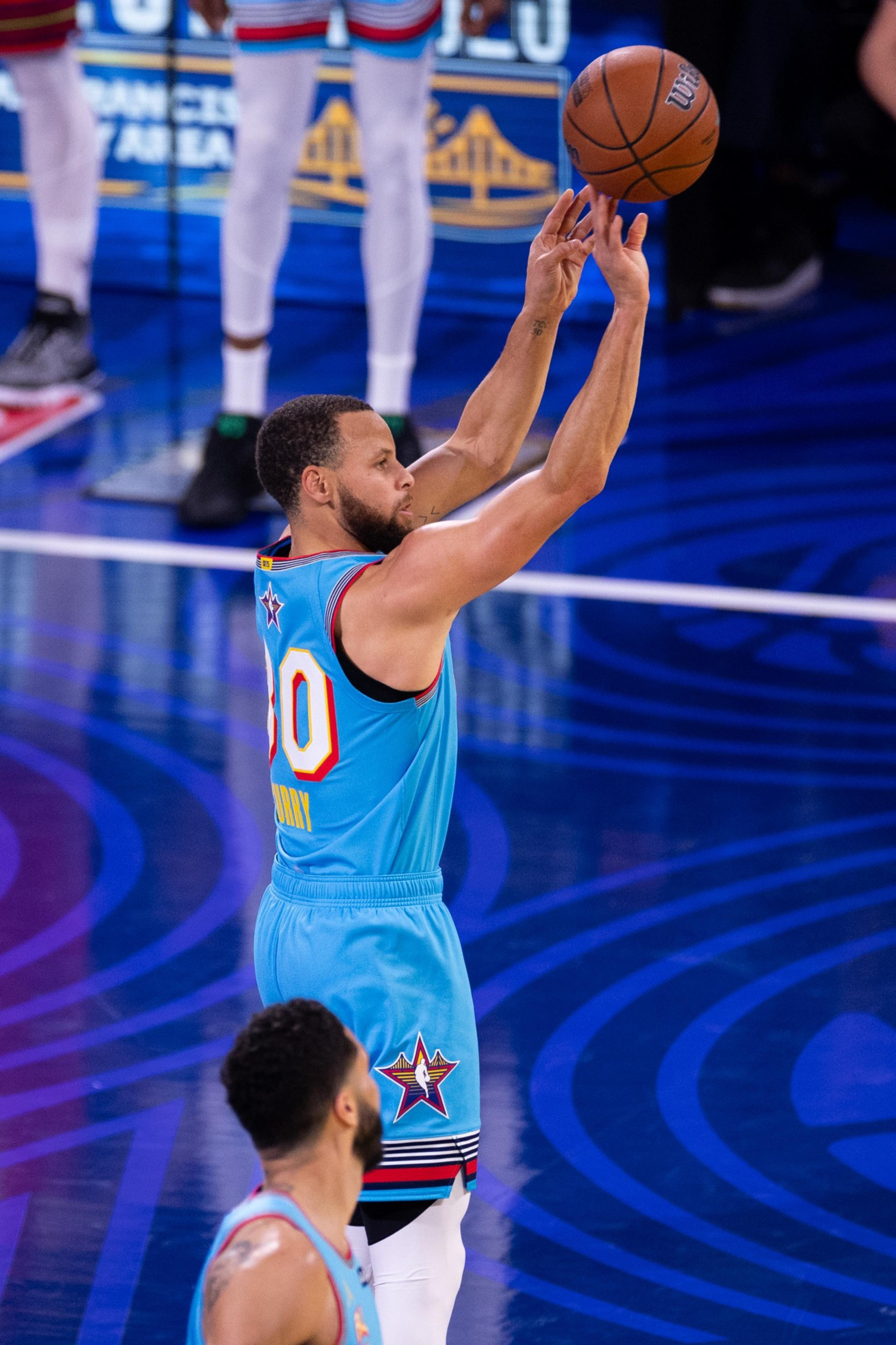 A basketball player in a blue jersey shoots a ball on a court. Another player is visible in the foreground, also wearing a similar blue jersey.