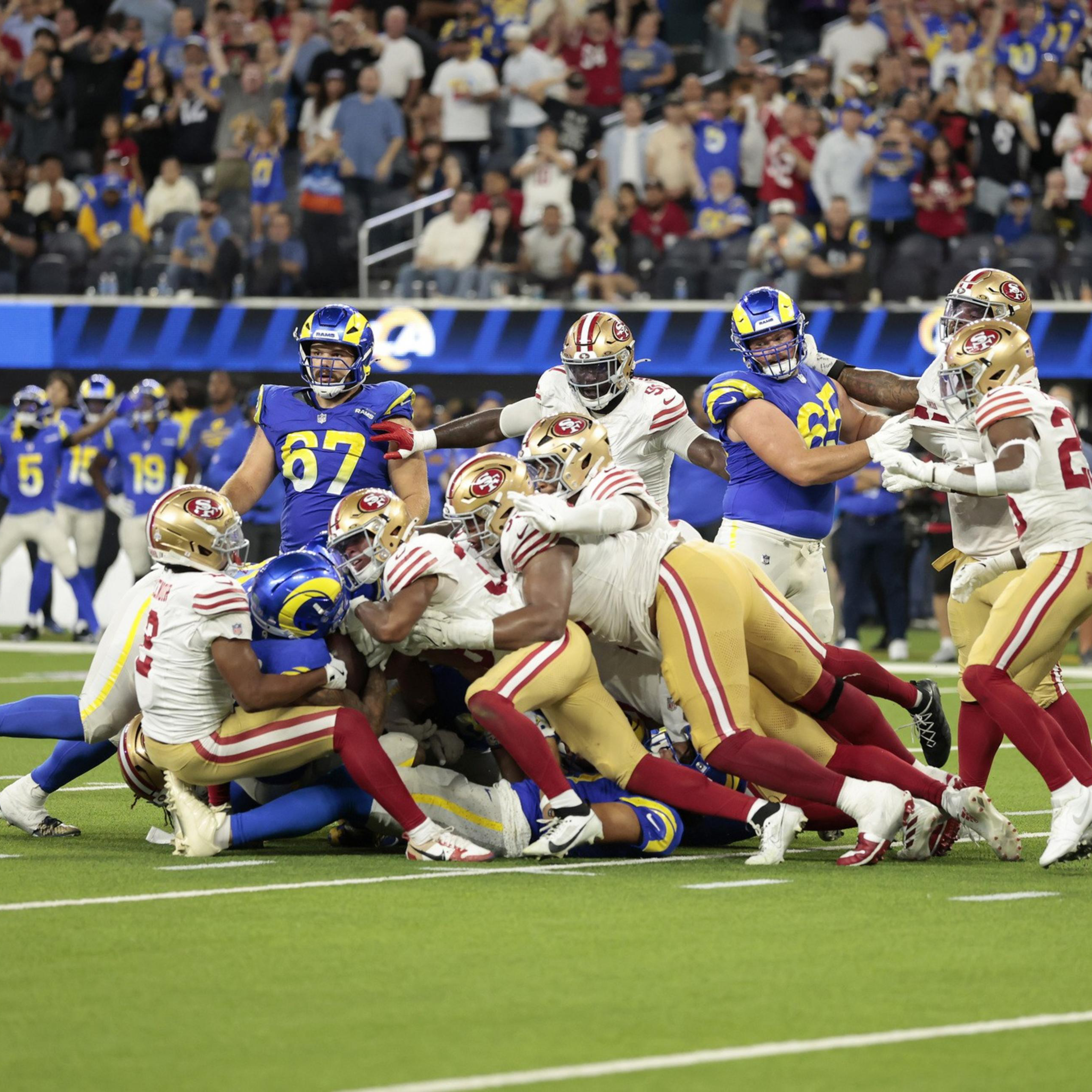 Several San Francisco 49ers players are tackling a Los Angeles Rams player on the football field during a crowded play.
