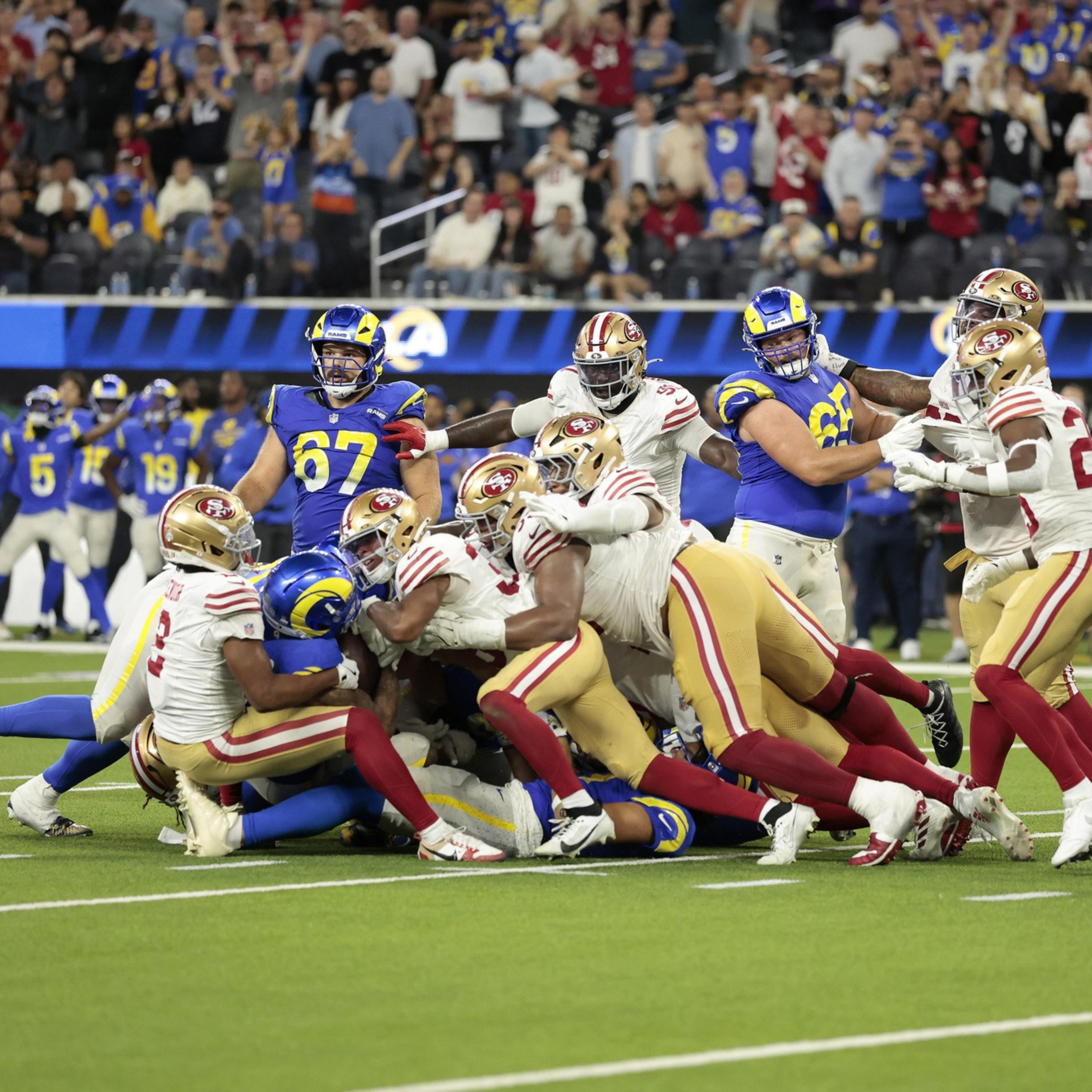 Several San Francisco 49ers players are tackling a Los Angeles Rams player on the football field during a crowded play.