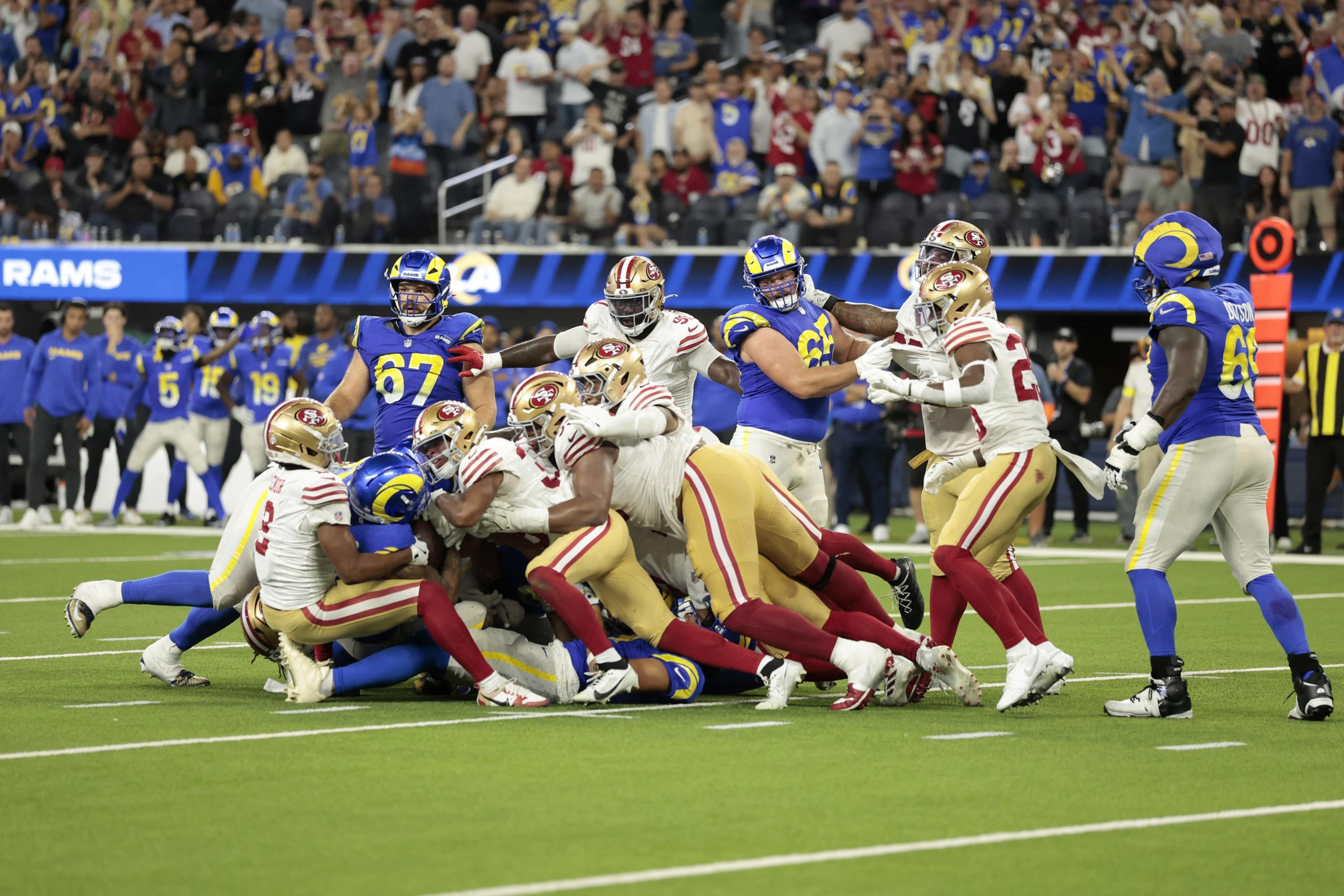 Several San Francisco 49ers players are tackling a Los Angeles Rams player on the football field during a crowded play.