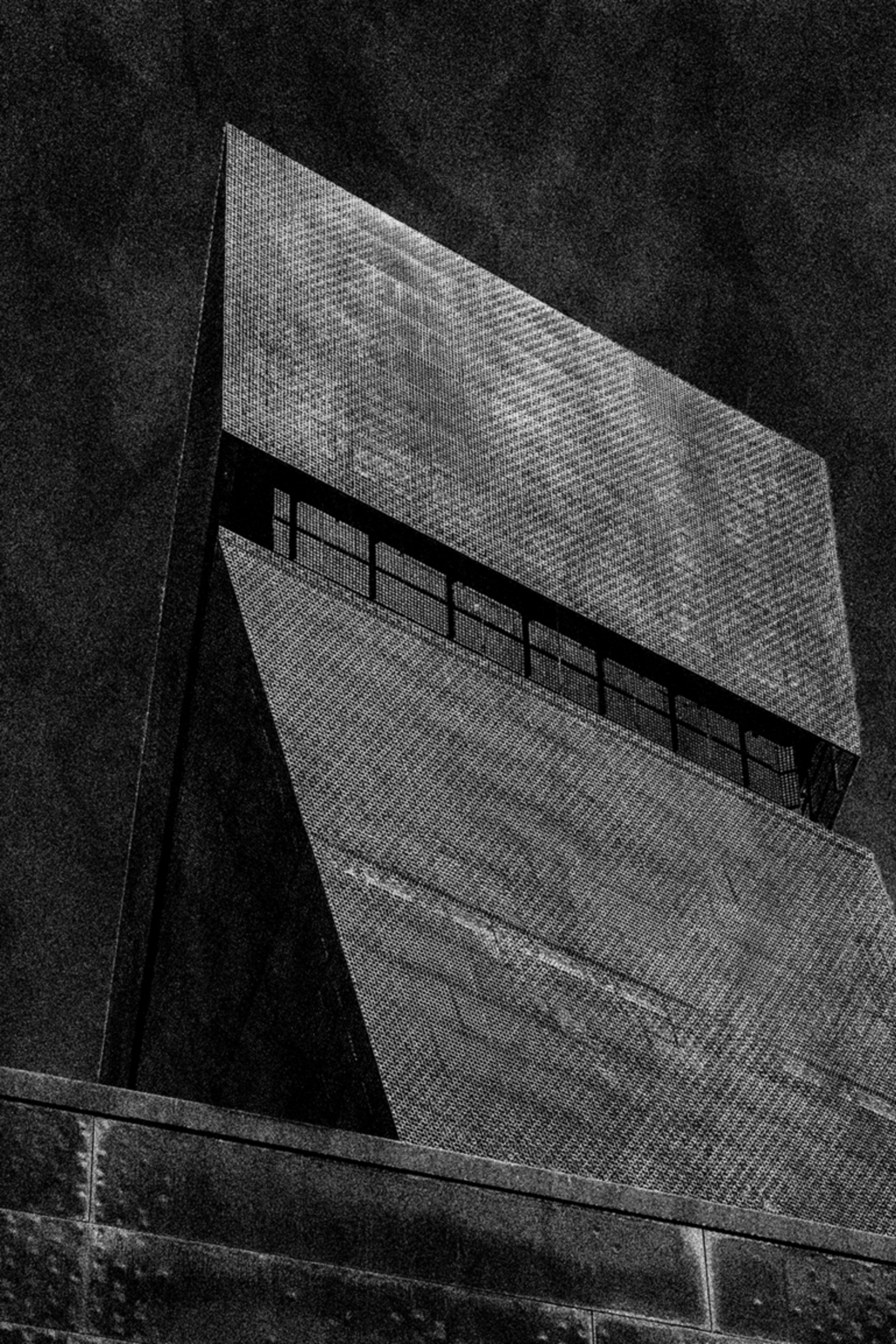 A shadow of a person holding a bag is cast on a brick wall next to a tall, angular concrete building, separated by a vertical yellow stripe.
