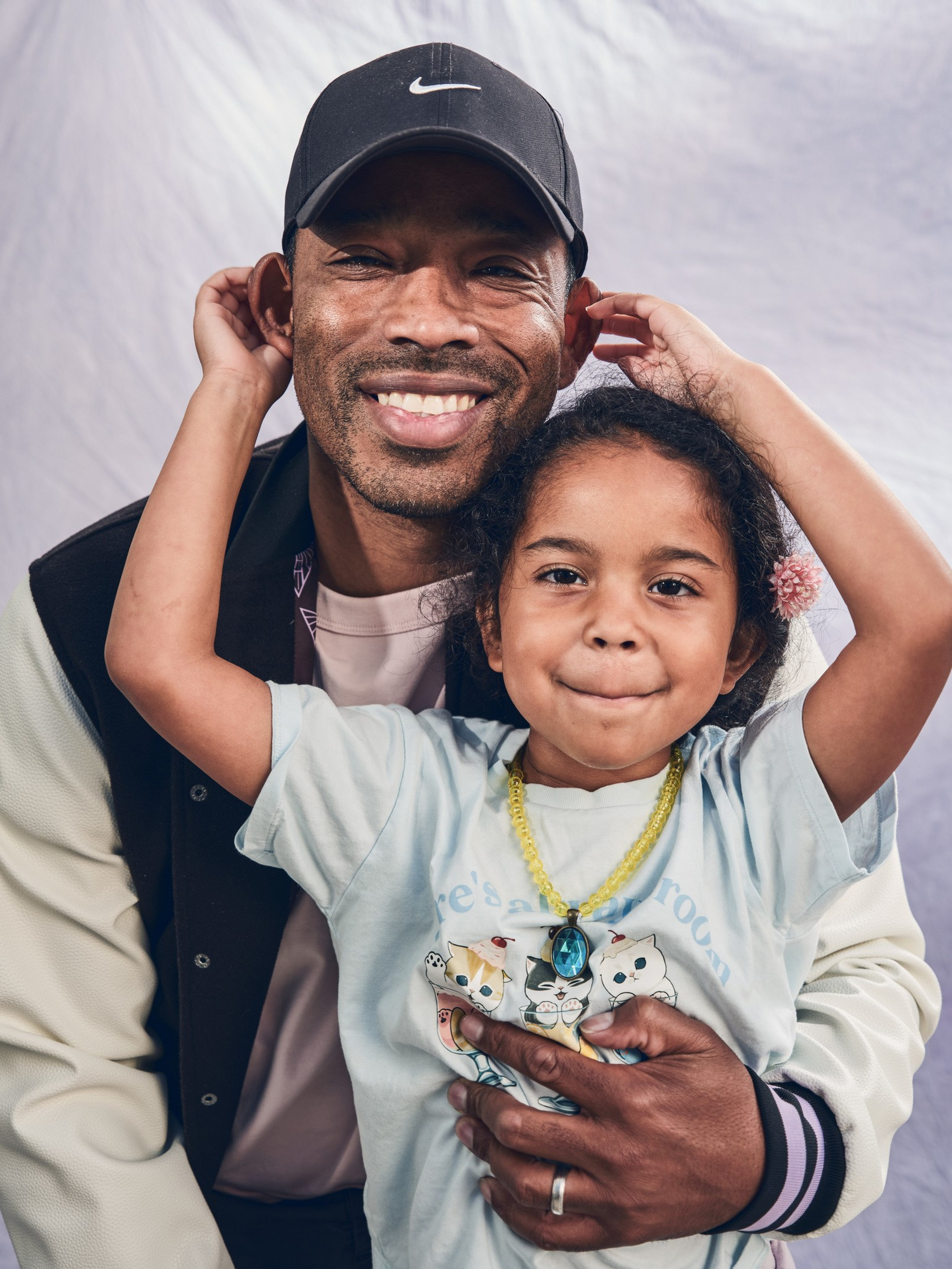 A smiling man wearing a black Nike cap hugs a young girl with curly hair, who playfully holds her ears up and wears a cat-themed shirt and necklace.