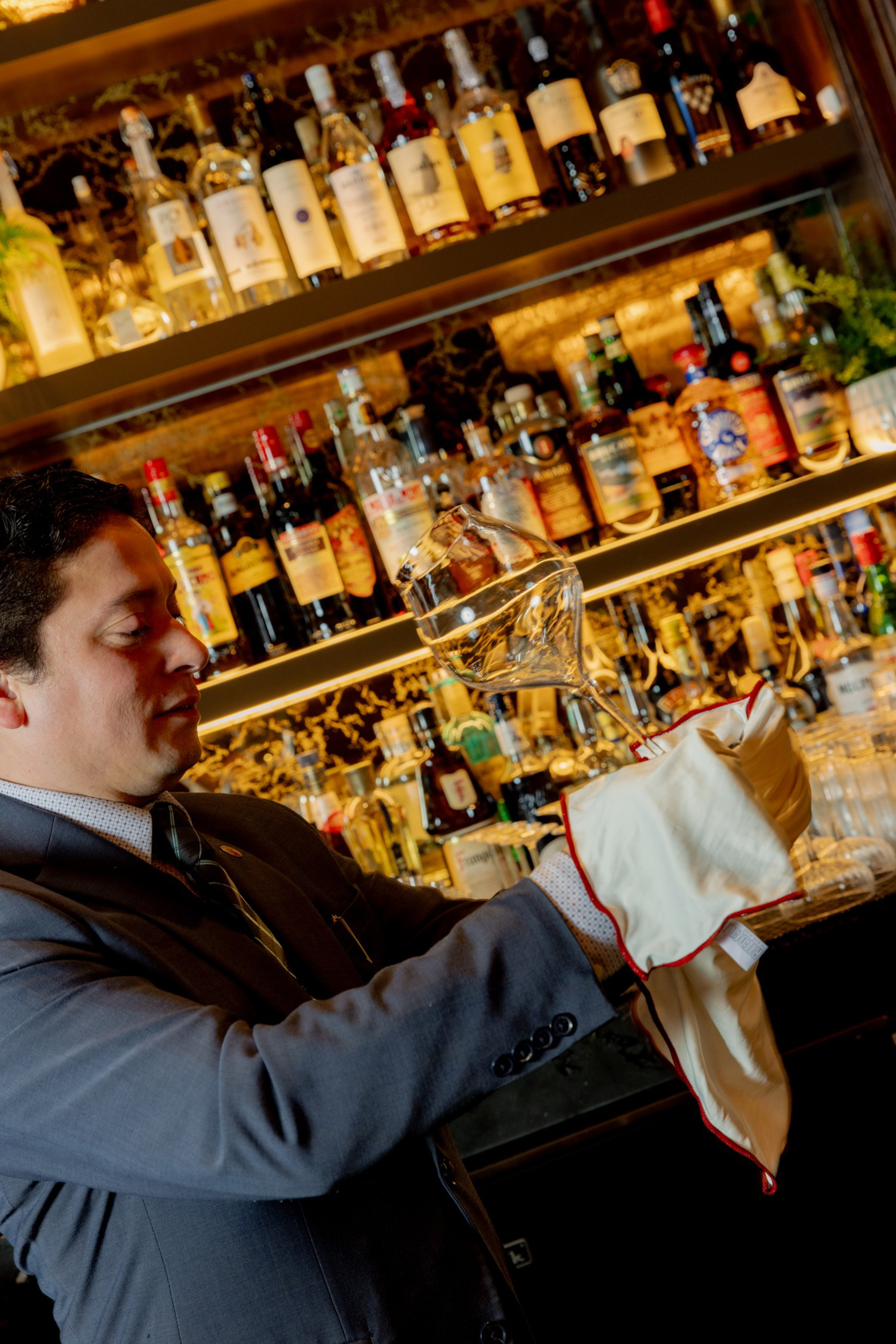 A bartender in a suit carefully polishes a wine glass with a white cloth in front of shelves filled with liquor bottles.
