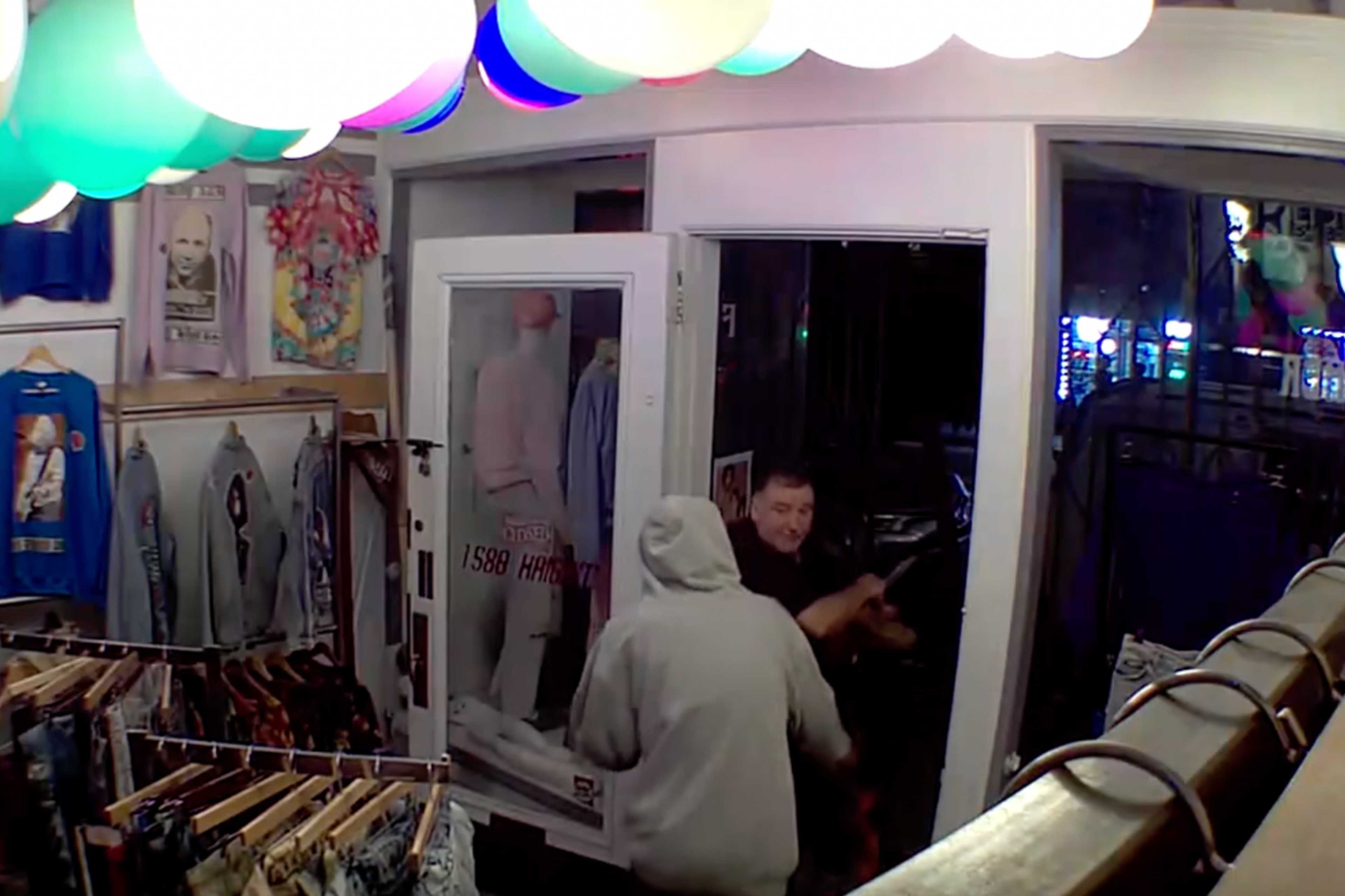 A person in a gray hoodie is inside a clothing store confronting another man near the open front door, surrounded by hanging clothes and colorful ceiling lights.