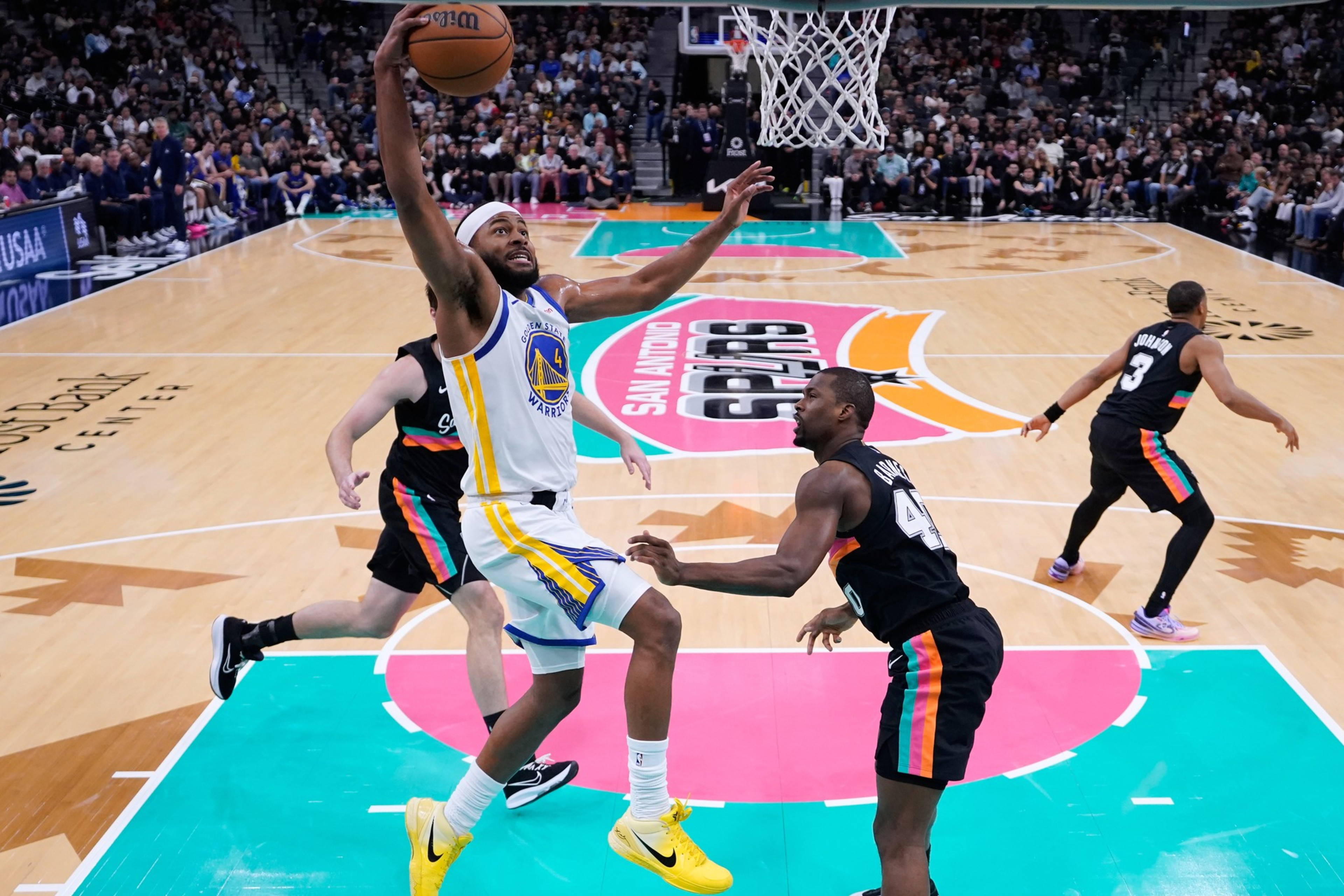 A basketball player in a white and blue uniform leaps toward the hoop for a shot while two defenders in black uniforms attempt to block him.