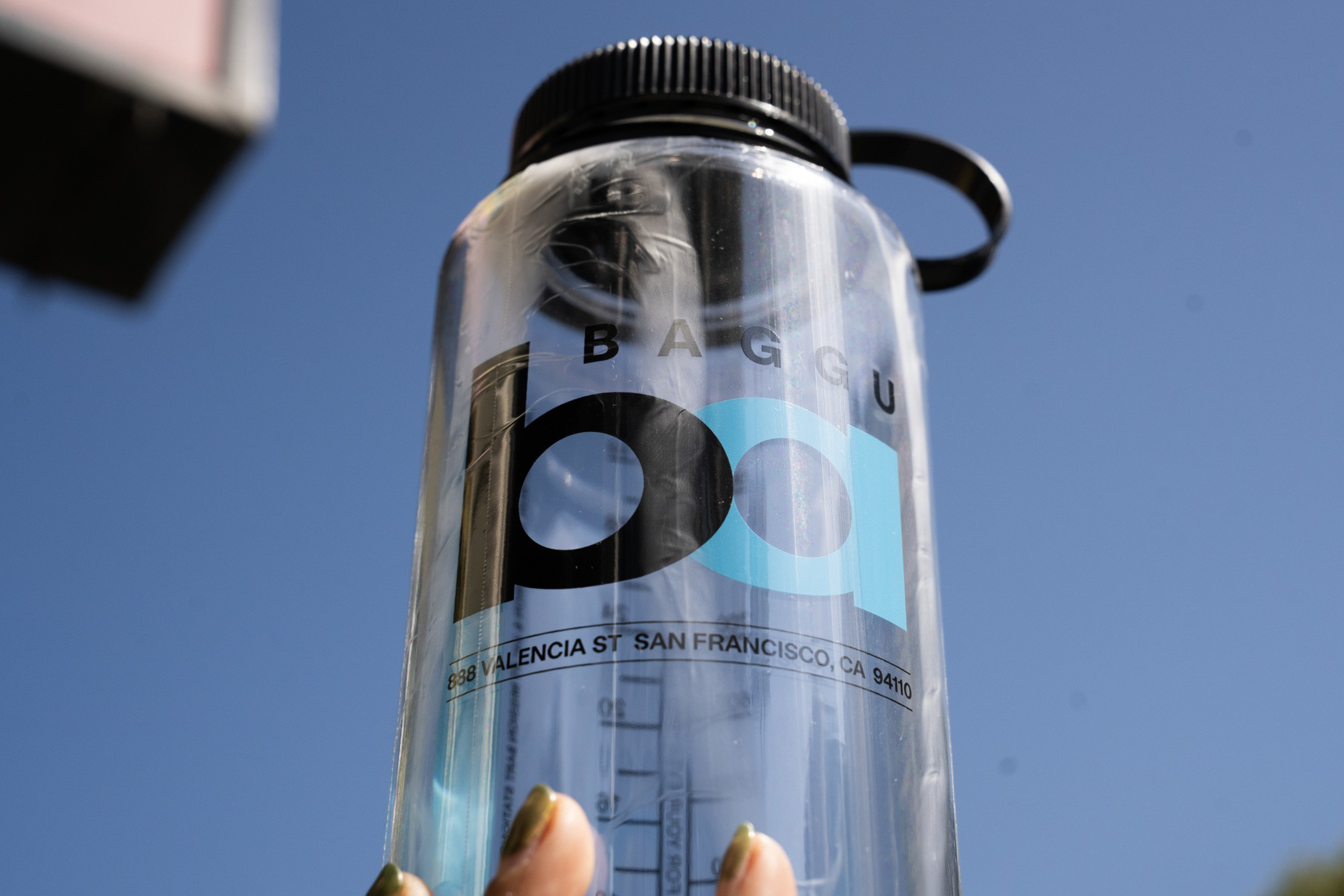 A clear plastic water bottle with a black lid and strap features the logo “BAGGU” and “100" along with an address in San Francisco.