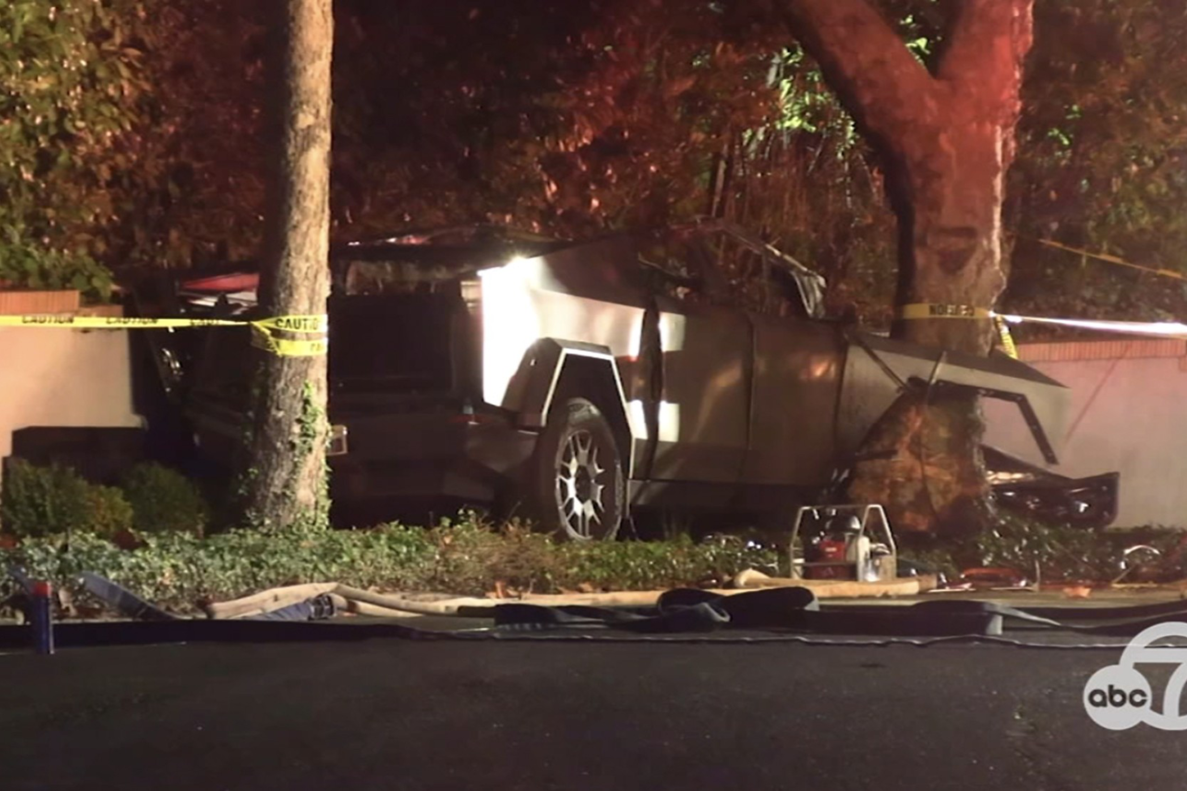 A damaged car is crashed into a tree beside a wall, surrounded by caution tape. It appears to be nighttime, with some debris on the ground.