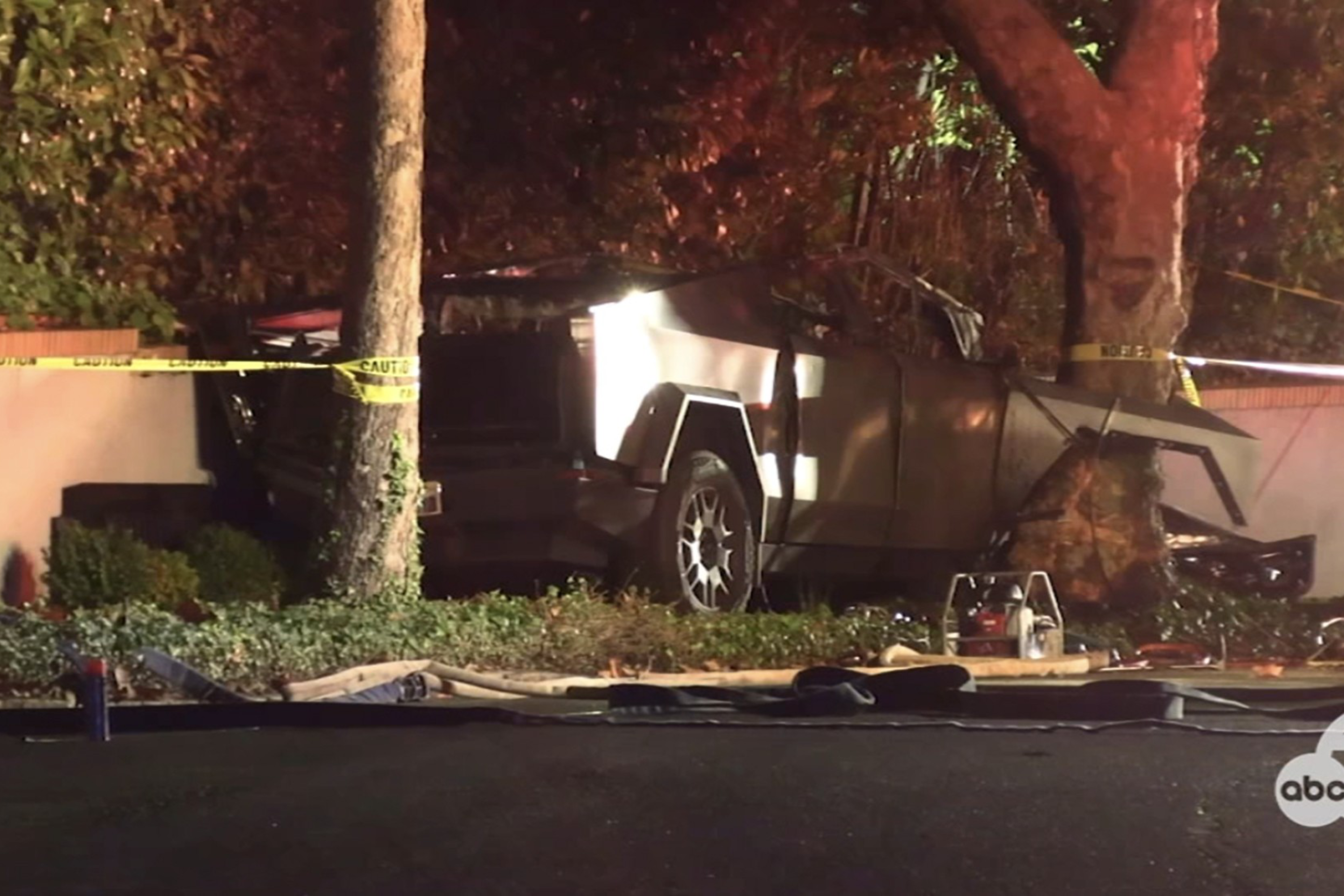 A damaged car is crashed into a tree beside a wall, surrounded by caution tape. It appears to be nighttime, with some debris on the ground.